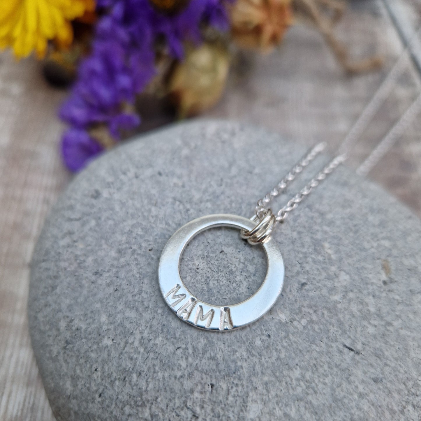 Sterling Silver ‘MAMA’ necklace. Silver disc with offset hole in centre, looks like a thick circle. Attached via two small silver hoops to silver chain. On thicker part of disc, inscription wording hammered into the silver reads MAMA in capitals. Disc measures approximately 20 mm in diameter. Chain length 18 inches.