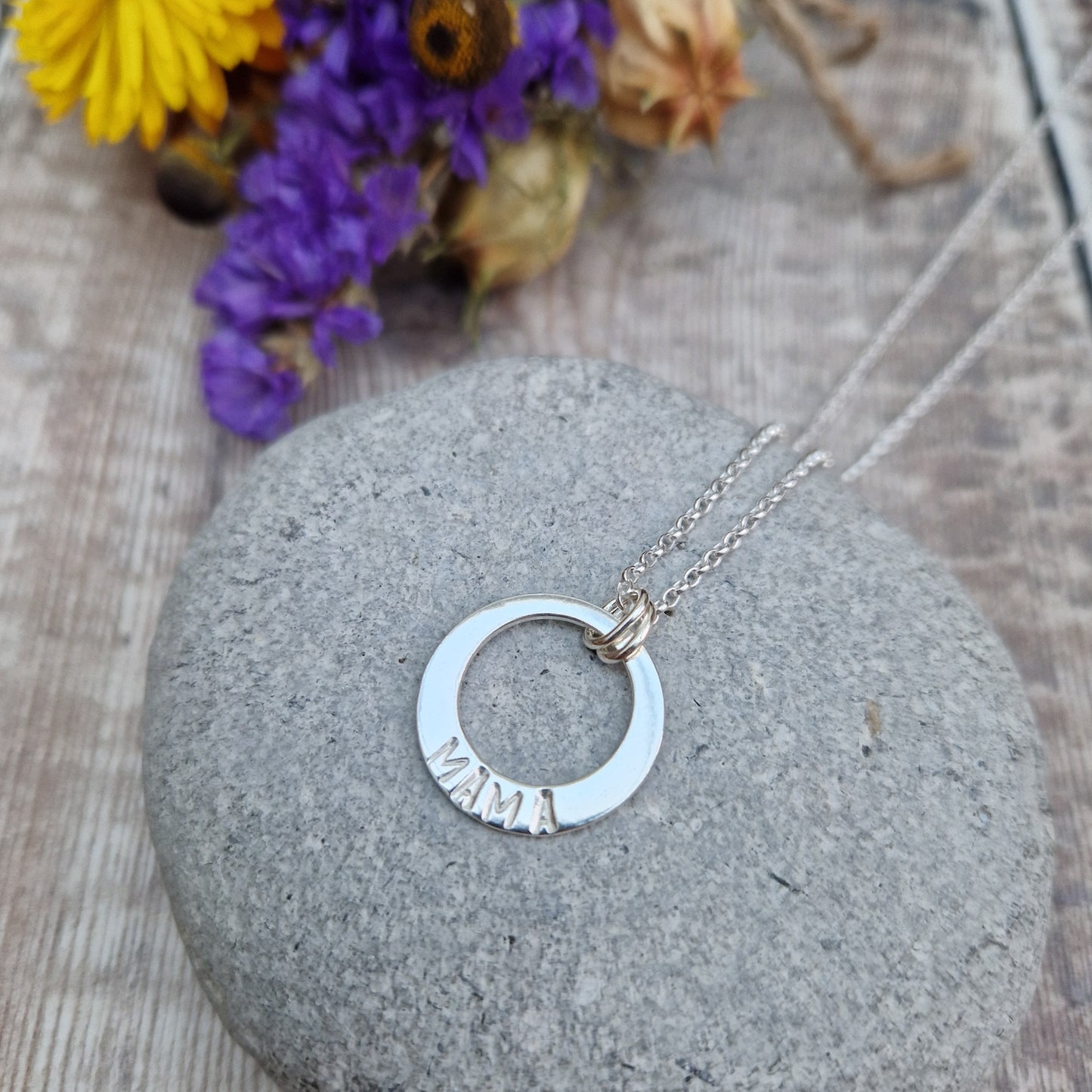 Sterling Silver ‘MAMA’ necklace. Silver disc with offset hole in centre, looks like a thick circle. Attached via two small silver hoops to silver chain. On thicker part of disc, inscription wording hammered into the silver reads MAMA in capitals. Disc measures approximately 20 mm in diameter. Chain length 18 inches.