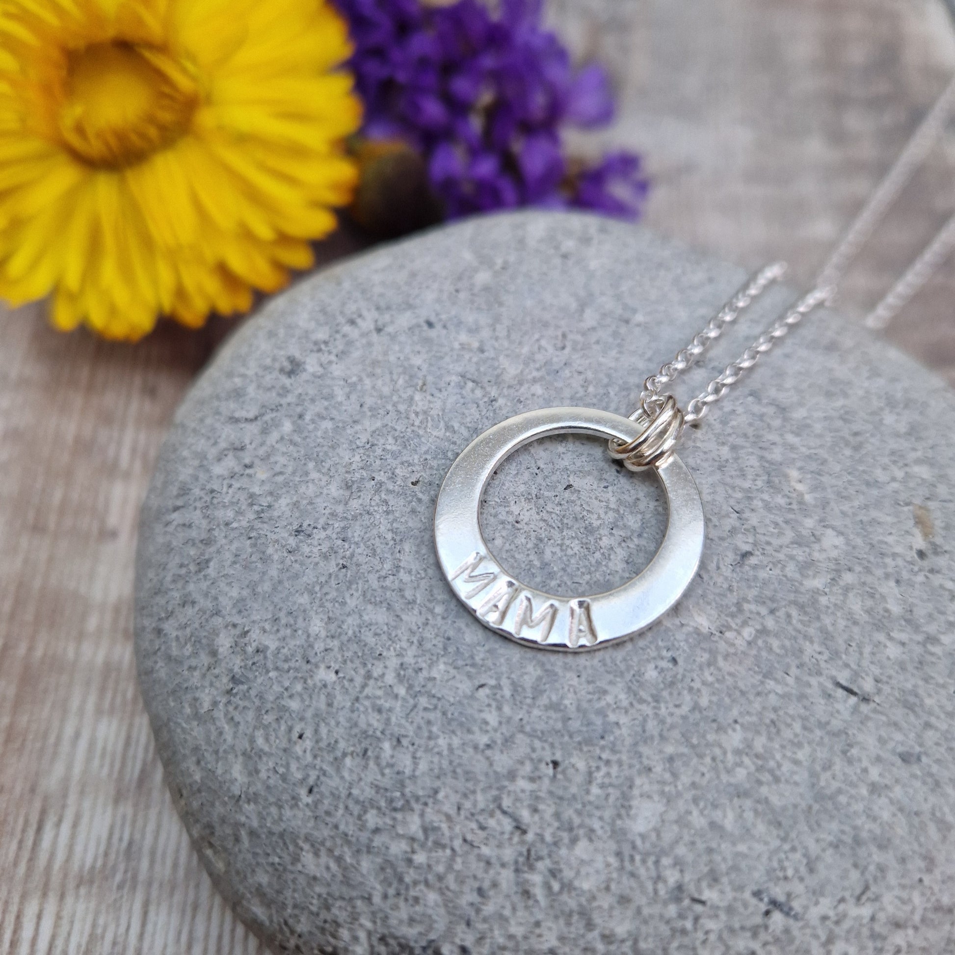 Sterling Silver ‘MAMA’ necklace. Silver disc with offset hole in centre, looks like a thick circle. Attached via two small silver hoops to silver chain. On thicker part of disc, inscription wording hammered into the silver reads MAMA in capitals. Disc measures approximately 20 mm in diameter. Chain length 18 inches.