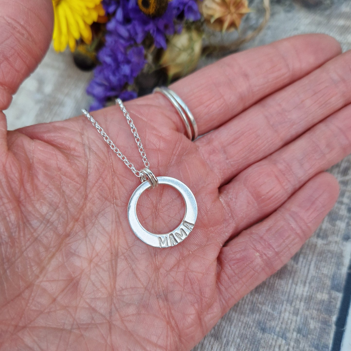 Sterling Silver ‘MAMA’ necklace. Silver disc with offset hole in centre, looks like a thick circle. Attached via two small silver hoops to silver chain. On thicker part of disc, inscription wording hammered into the silver reads MAMA in capitals. Disc measures approximately 20 mm in diameter. Chain length 18 inches.