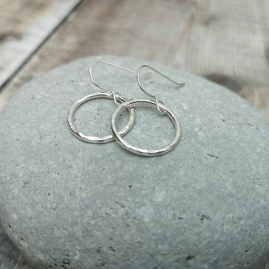 A close-up of medium-sized sterling silver hammered circle earrings resting on a smooth grey stone. The subtle texture of the circles reflects light, enhancing their minimalist and handcrafted design.