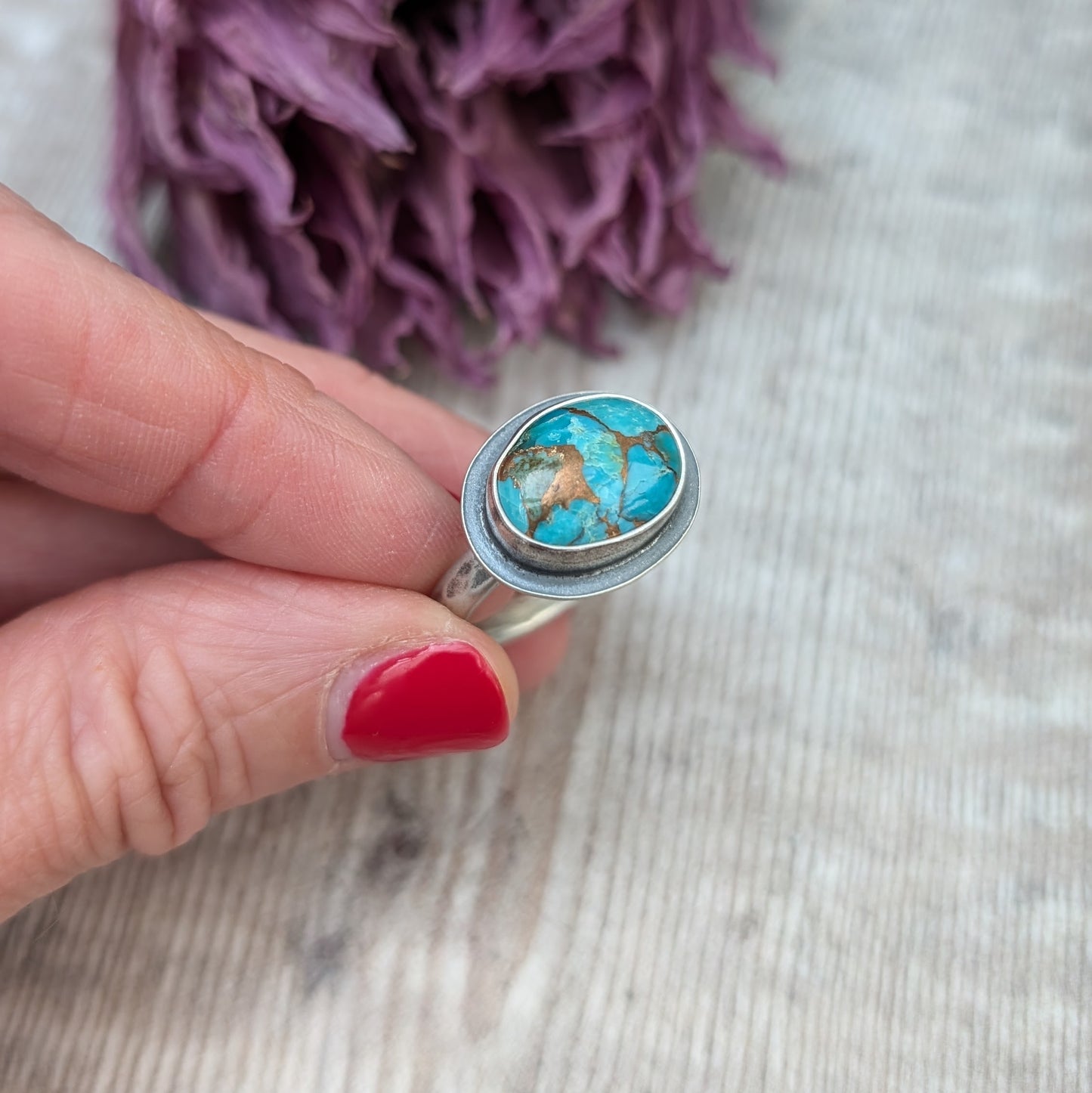 A hand with red painted nails holds a silver ring with a Mohave Turquoise Stone against a backdrop of dried purple flowers.