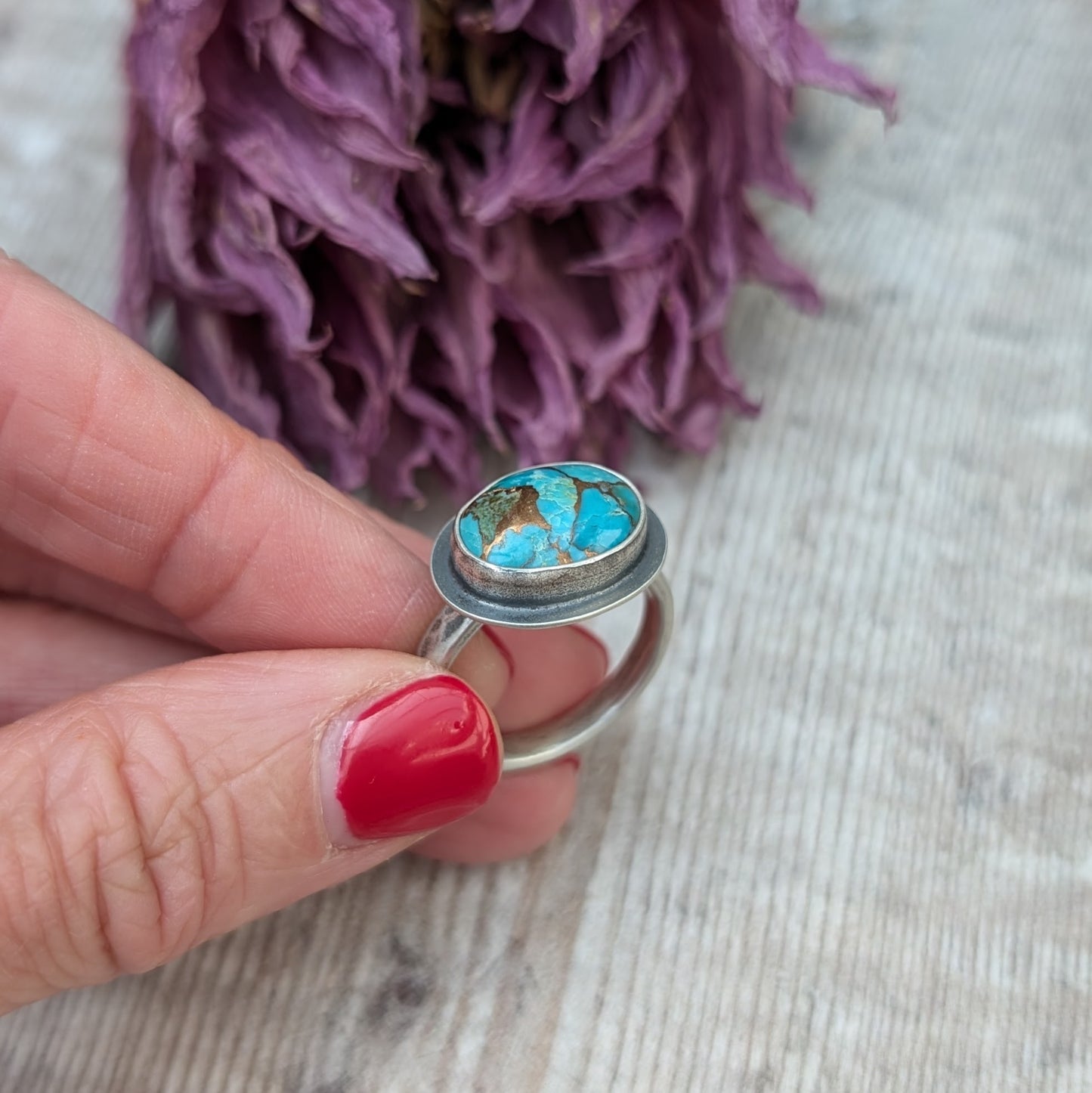 A hand with red painted nails holds a silver ring with a Mohave Turquoise Stone against a backdrop of dried purple flowers.