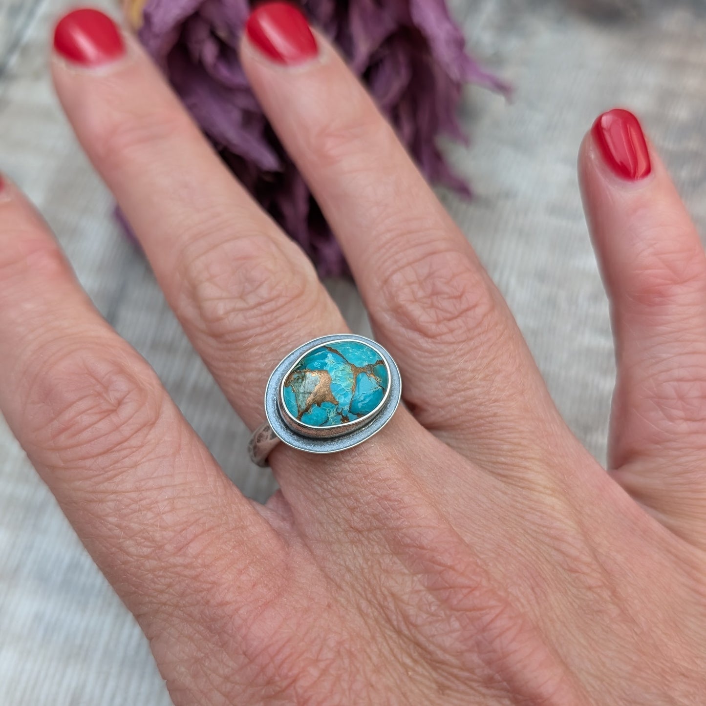 A hand with red painted nails holds a silver ring with a Mohave Turquoise Stone against a backdrop of dried purple flowers.