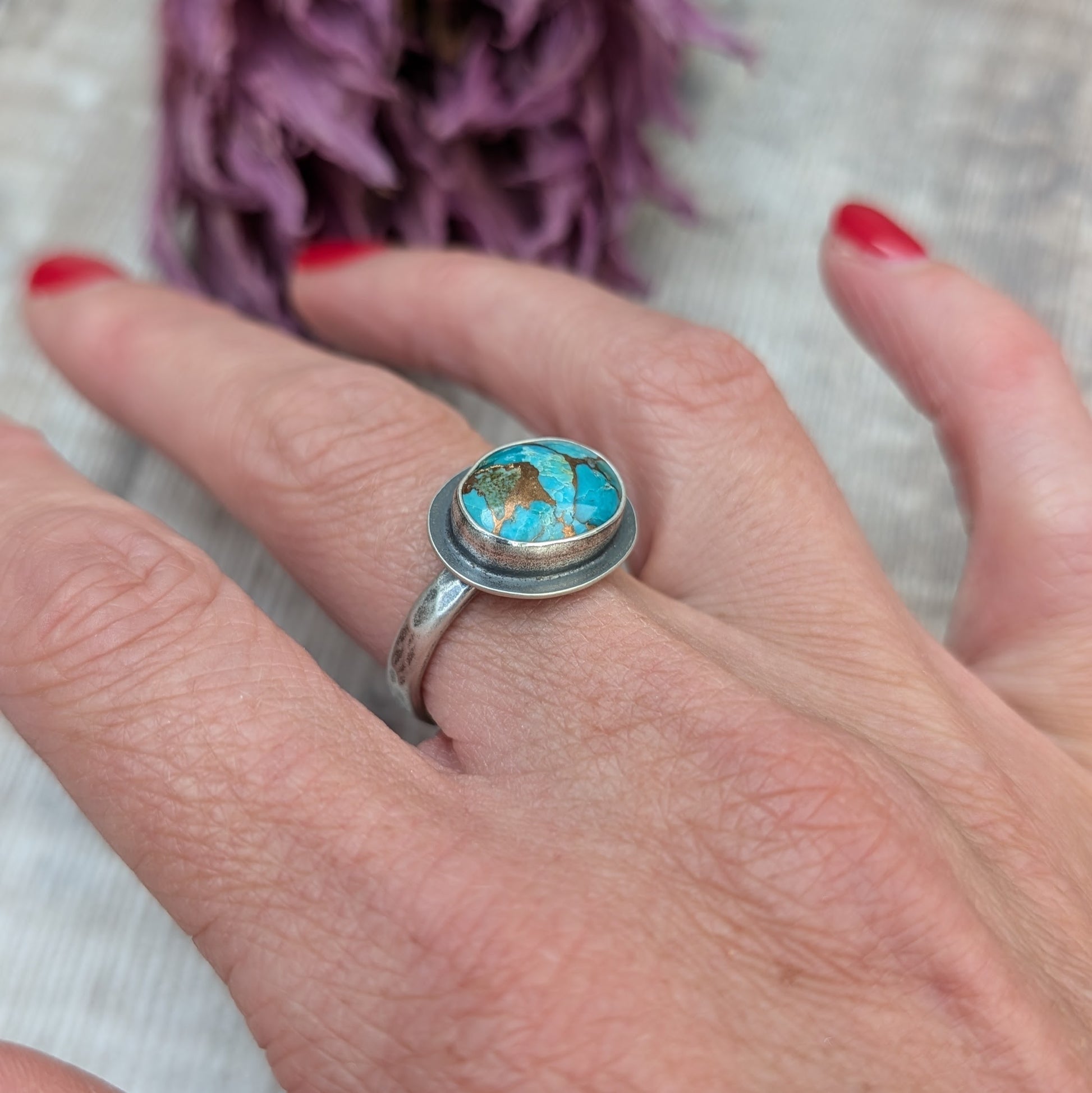 A hand with red painted nails holds a silver ring with a Mohave Turquoise Stone against a backdrop of dried purple flowers.