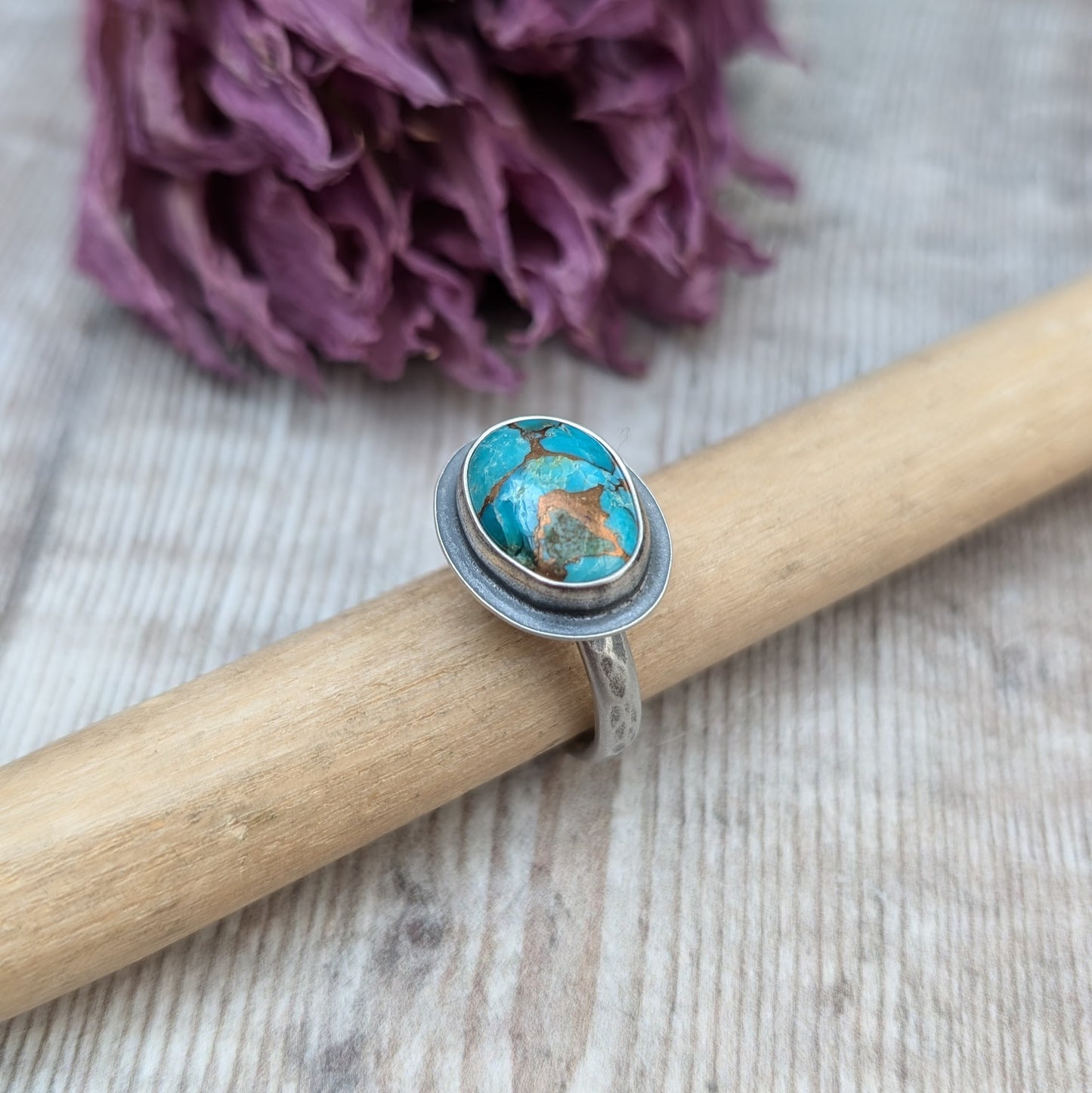 A silver ring with a Mohave Turquoise gemstone propped up on a piece of wood against a backdrop of dried purple flowers.