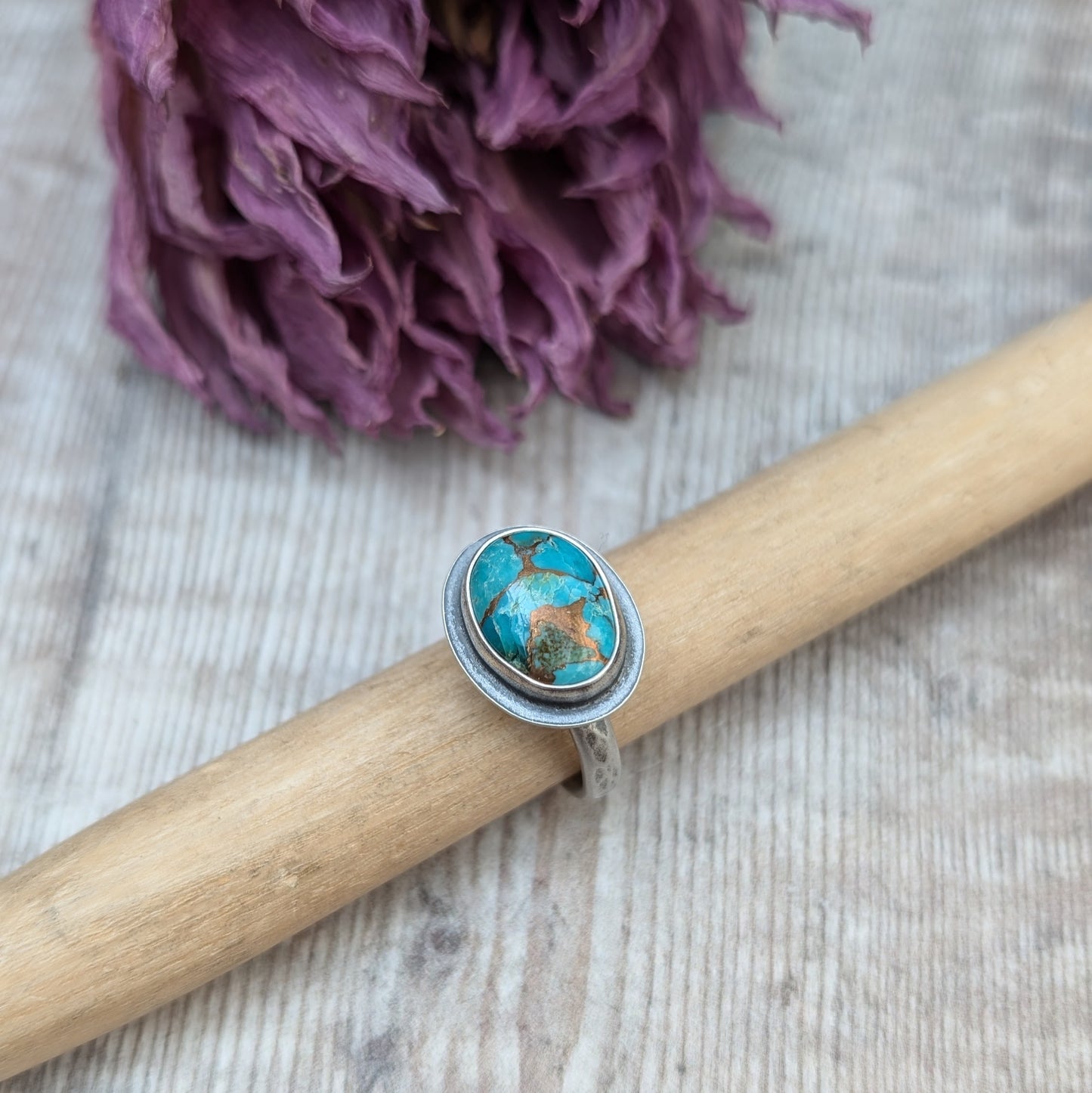 A silver ring with a Mohave Turquoise gemstone propped up on a piece of wood against a backdrop of dried purple flowers.