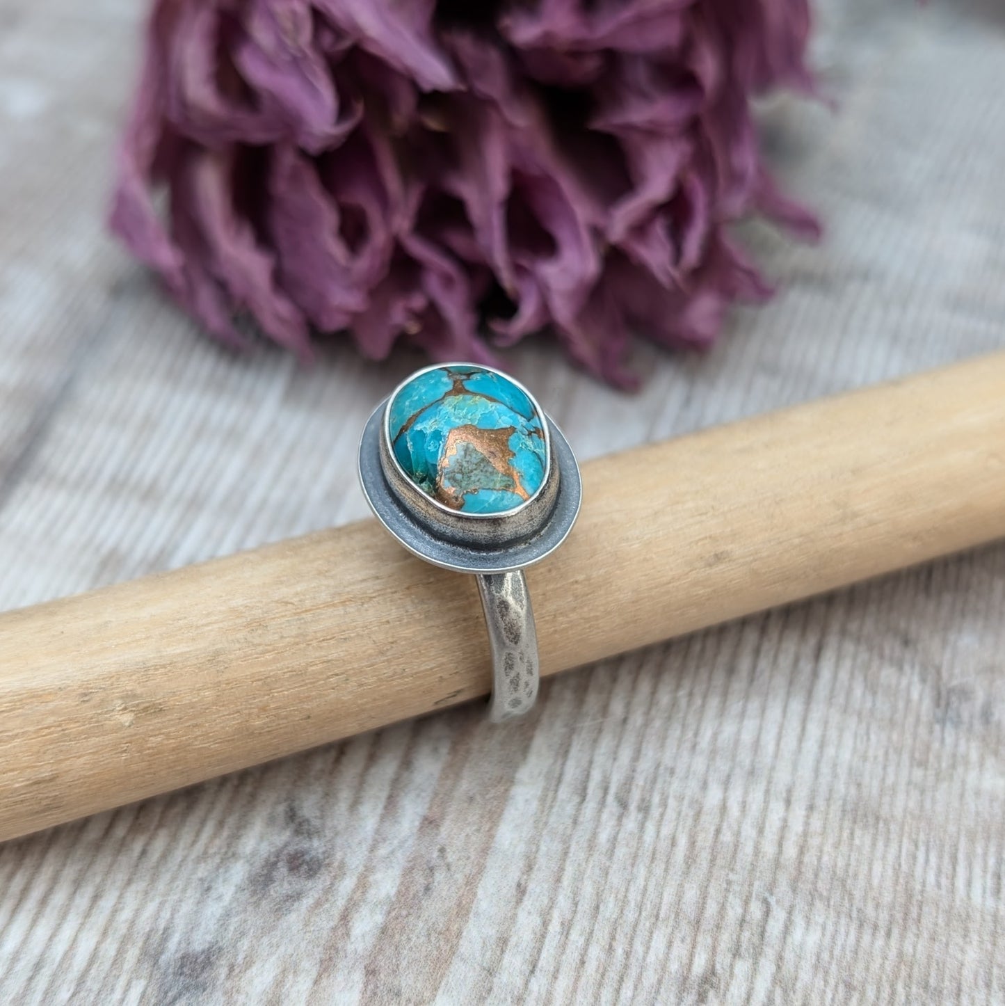 A silver ring with a Mohave Turquoise gemstone propped up on a piece of wood against a backdrop of dried purple flowers.