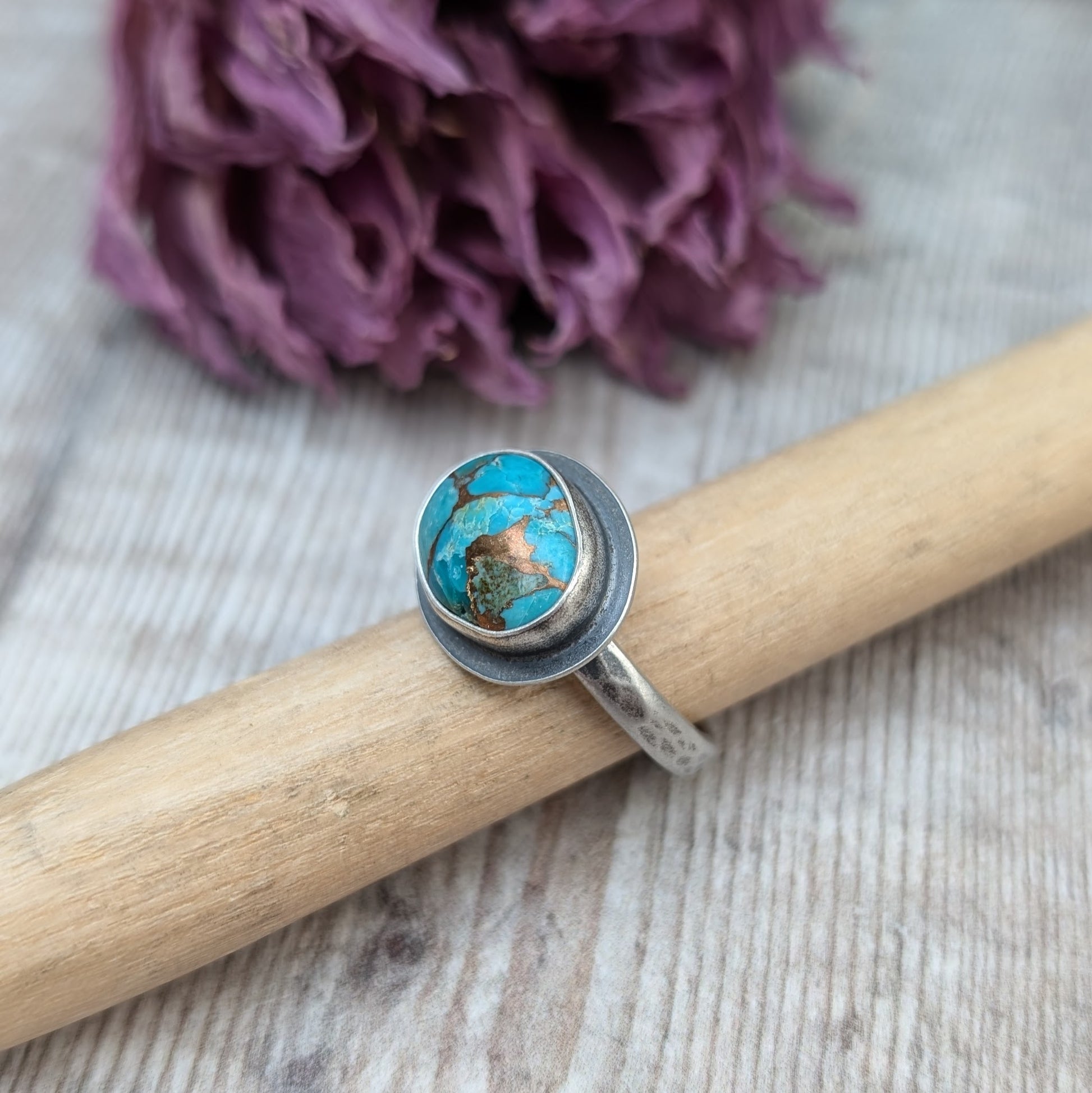 A silver ring with a Mohave Turquoise gemstone propped up on a piece of wood against a backdrop of dried purple flowers.