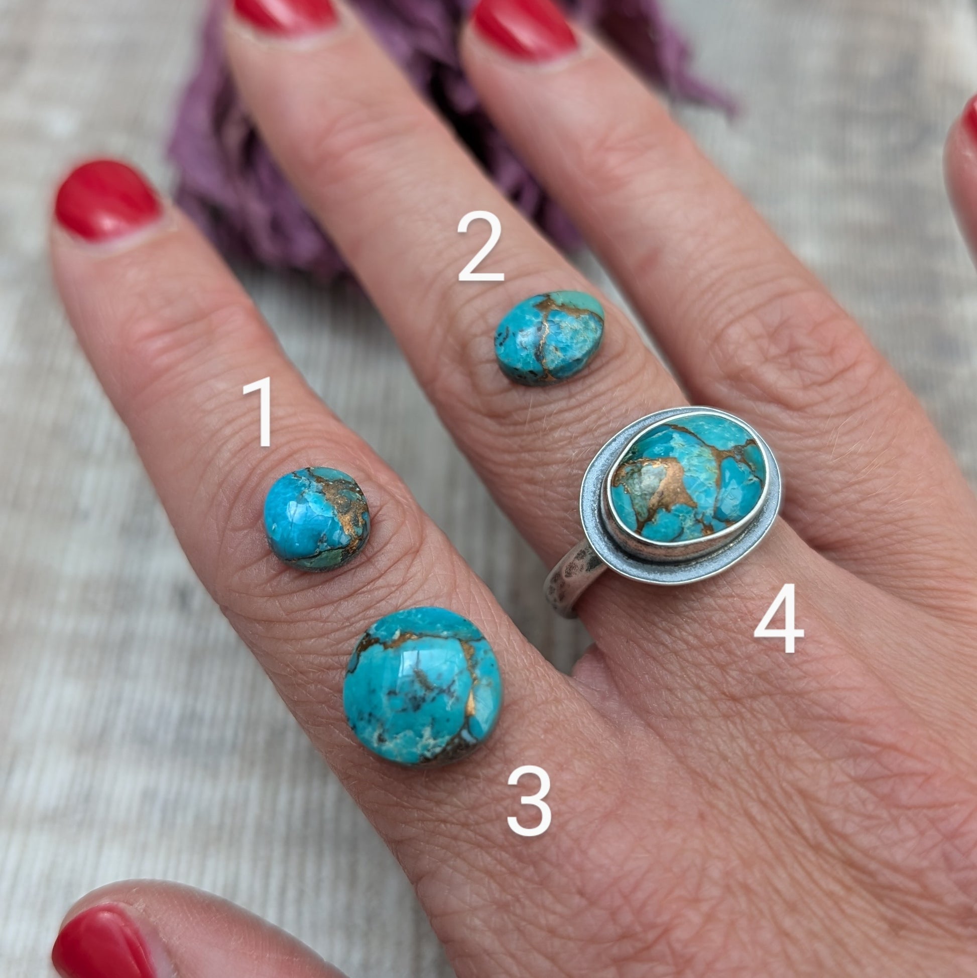 A hand with red painted nails holds a silver ring with a Mohave Turquoise Stone and a selection of stones against a backdrop of dried purple flowers.