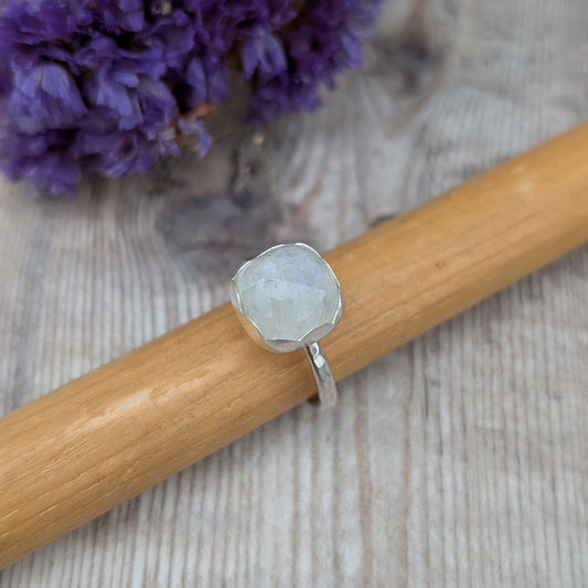 Silver Moonstone Ring displayed on a wooden stick with a blurred background, focusing on the round moonstone and hammered silver band.