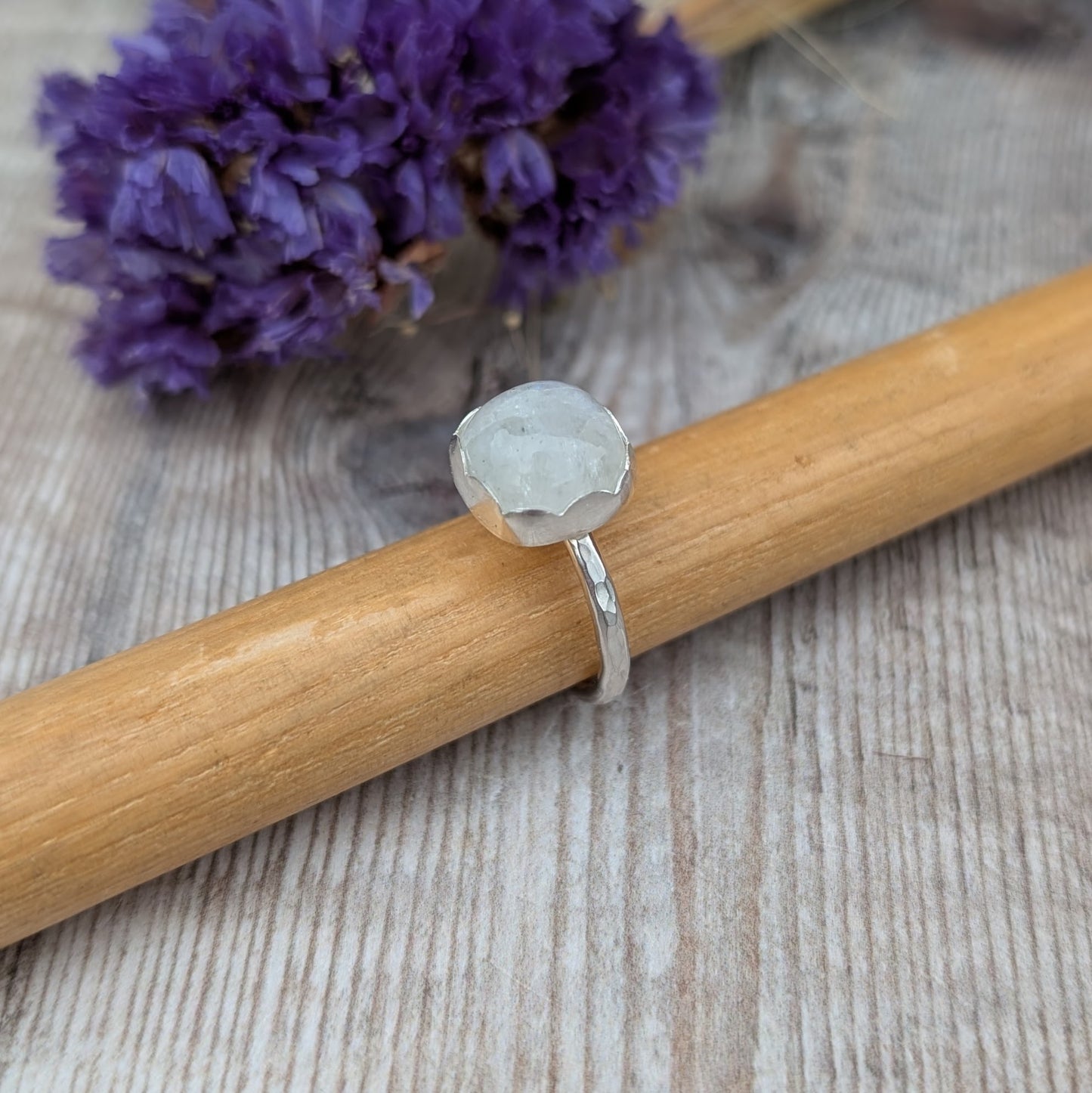 Silver Moonstone Ring resting on a wooden stick with purple flowers in the background, showing the hammered silver band and round moonstone.