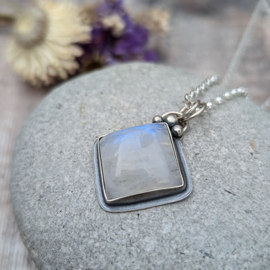 Close-up of a square-cut moonstone necklace on a grey stone background. The moonstone is set in oxidised recycled sterling silver, with silver bead details, and shows a gentle blue iridescence.