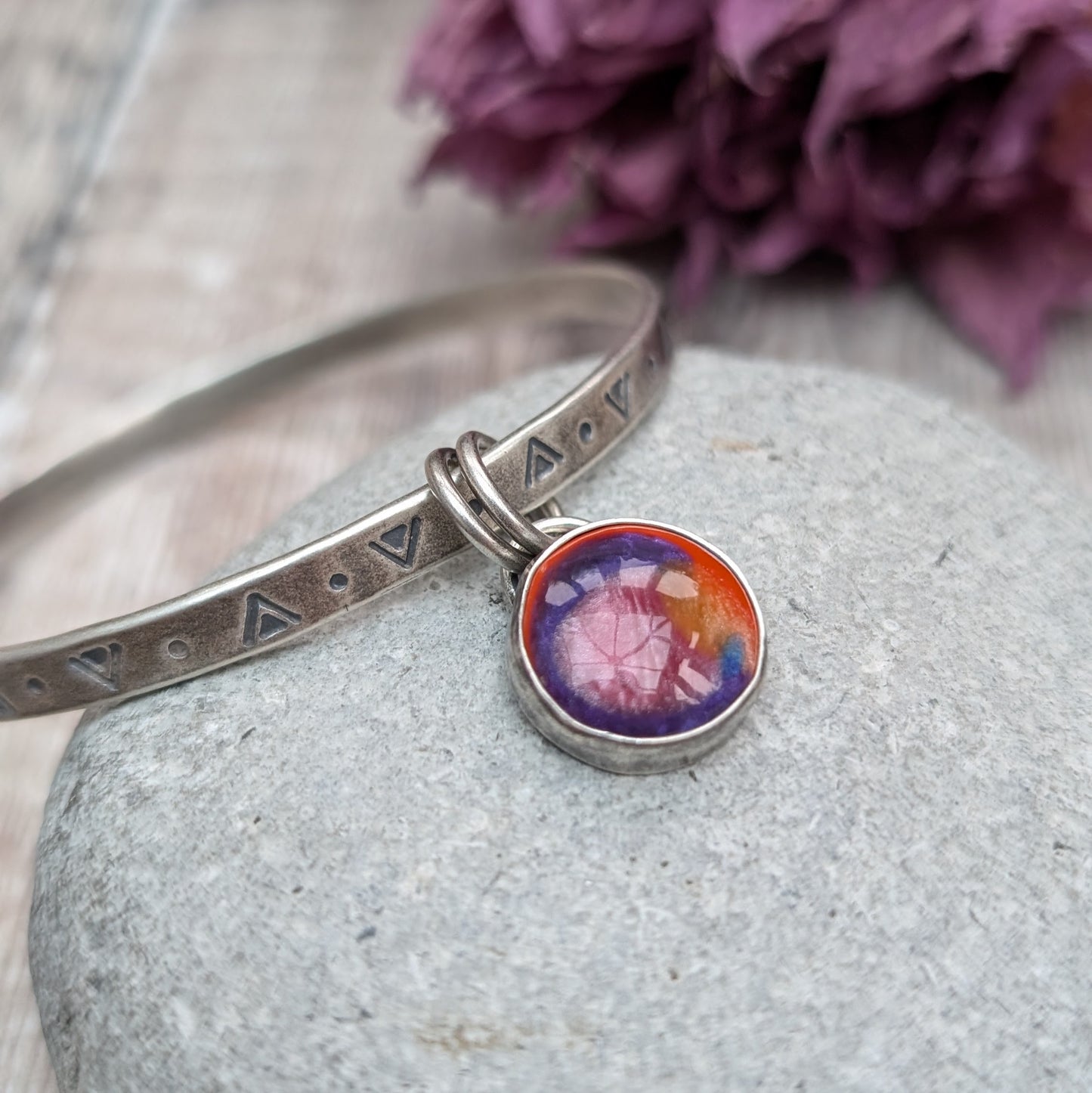 A silver bangle with triangular and circular etchings is placed on a smooth, grey stone. The bangle features a round, multicolored charm with shades of red, orange, and purple, which hangs from two small rings.