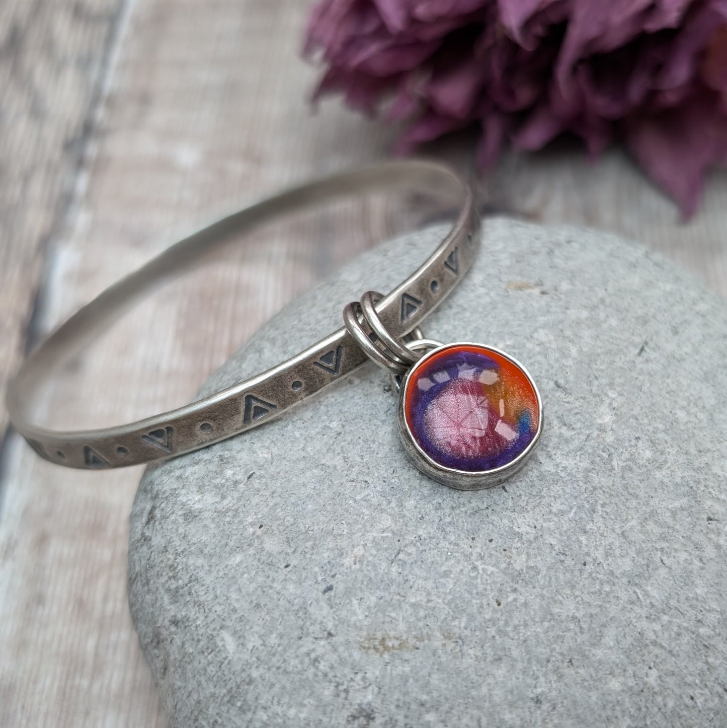 A silver bangle with triangular and circular etchings is placed on a smooth, grey stone. The bangle features a round, multicolored charm with shades of red, orange, and purple, which hangs from two small rings.