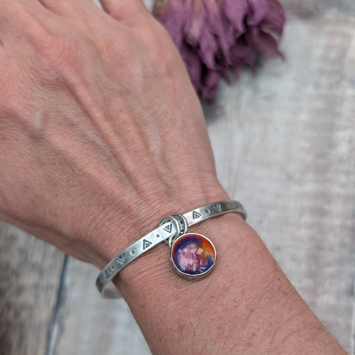 A close-up of a person's wrist wearing a silver bangle a silver bangle with triangular and circular etchings. The bangle features a round, multicolored charm with shades of red, orange, and purple, which hangs from two small rings.