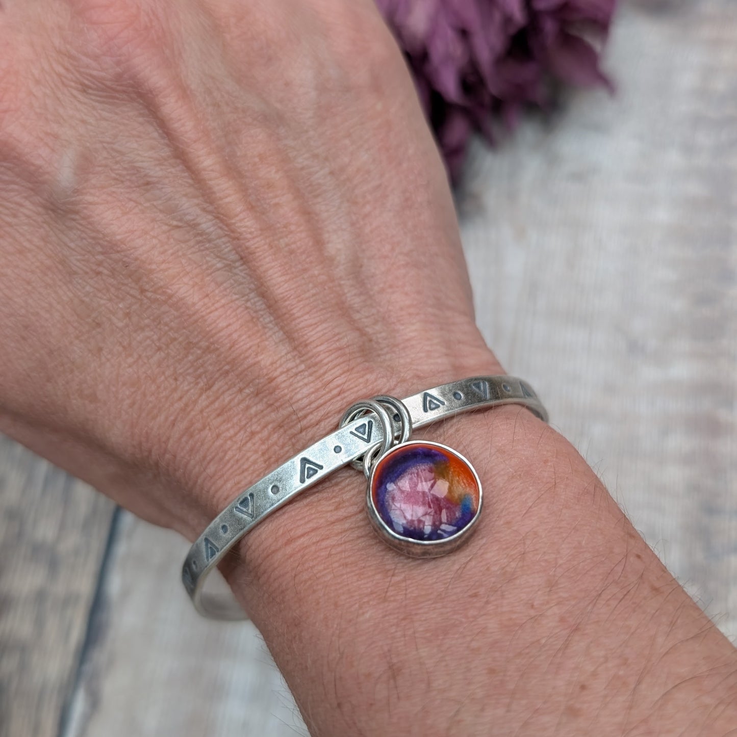 A close-up of a person's wrist wearing a silver bangle a silver bangle with triangular and circular etchings. The bangle features a round, multicolored charm with shades of red, orange, and purple, which hangs from two small rings.