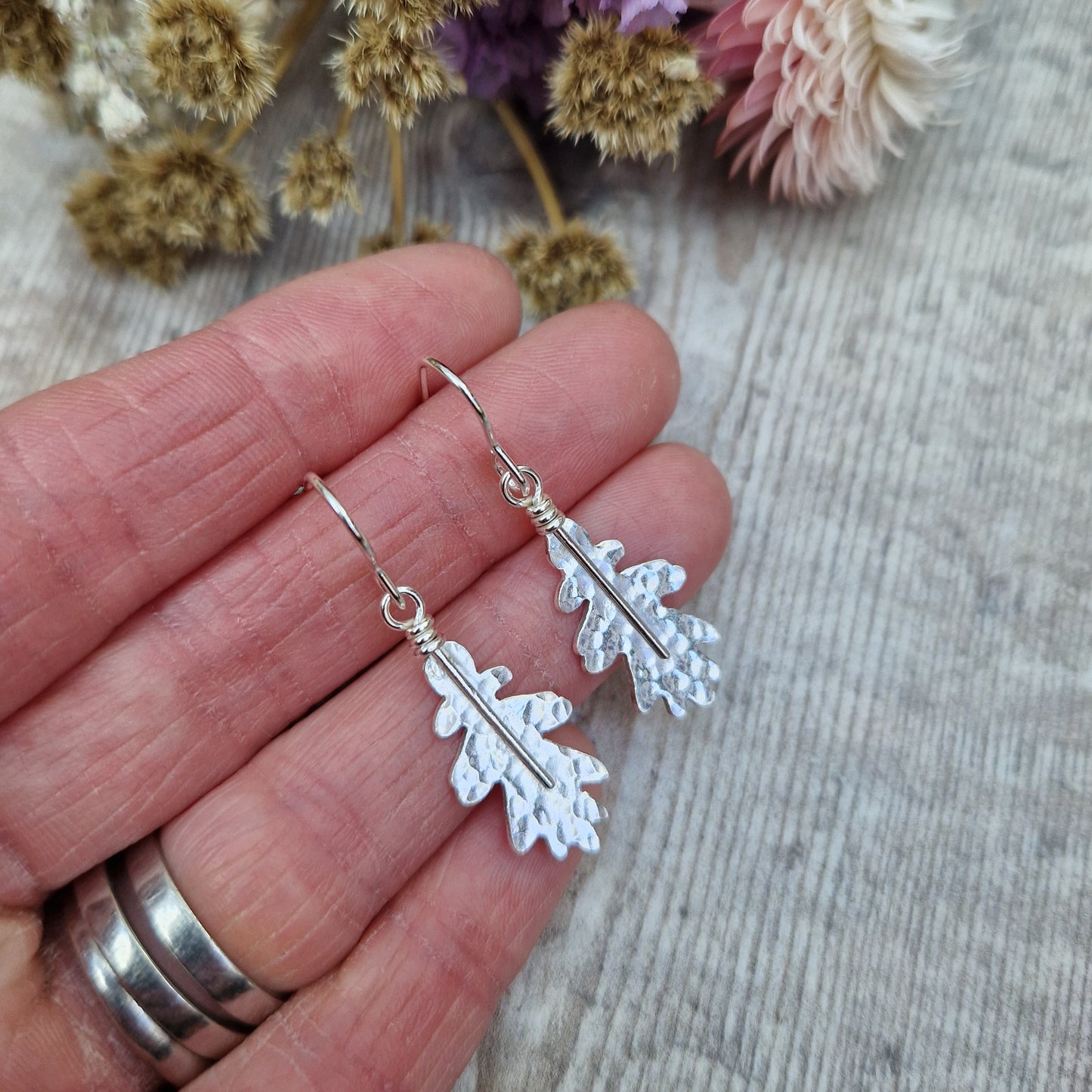 A pair of sterling silver oak leaf earrings held in hand, showcasing their delicate hammered texture. The earrings dangle from simple silver hooks, with dried flowers in the background.