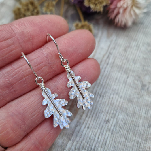 A pair of sterling silver oak leaf earrings held in hand, showcasing their delicate hammered texture. The earrings dangle from simple silver hooks, with dried flowers in the background.