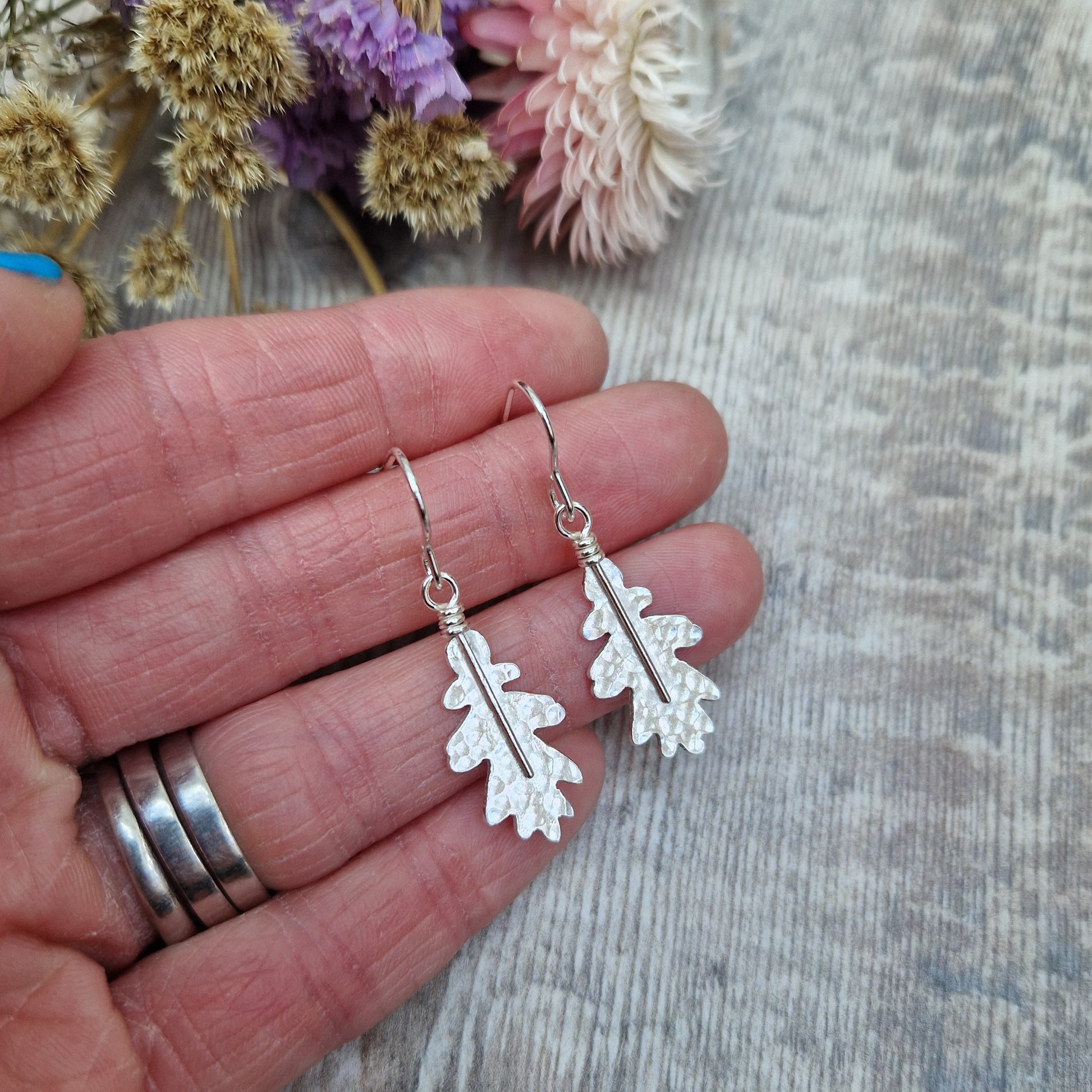 A pair of sterling silver oak leaf earrings held in hand, showcasing their delicate hammered texture. The earrings dangle from simple silver hooks, with dried flowers in the background.