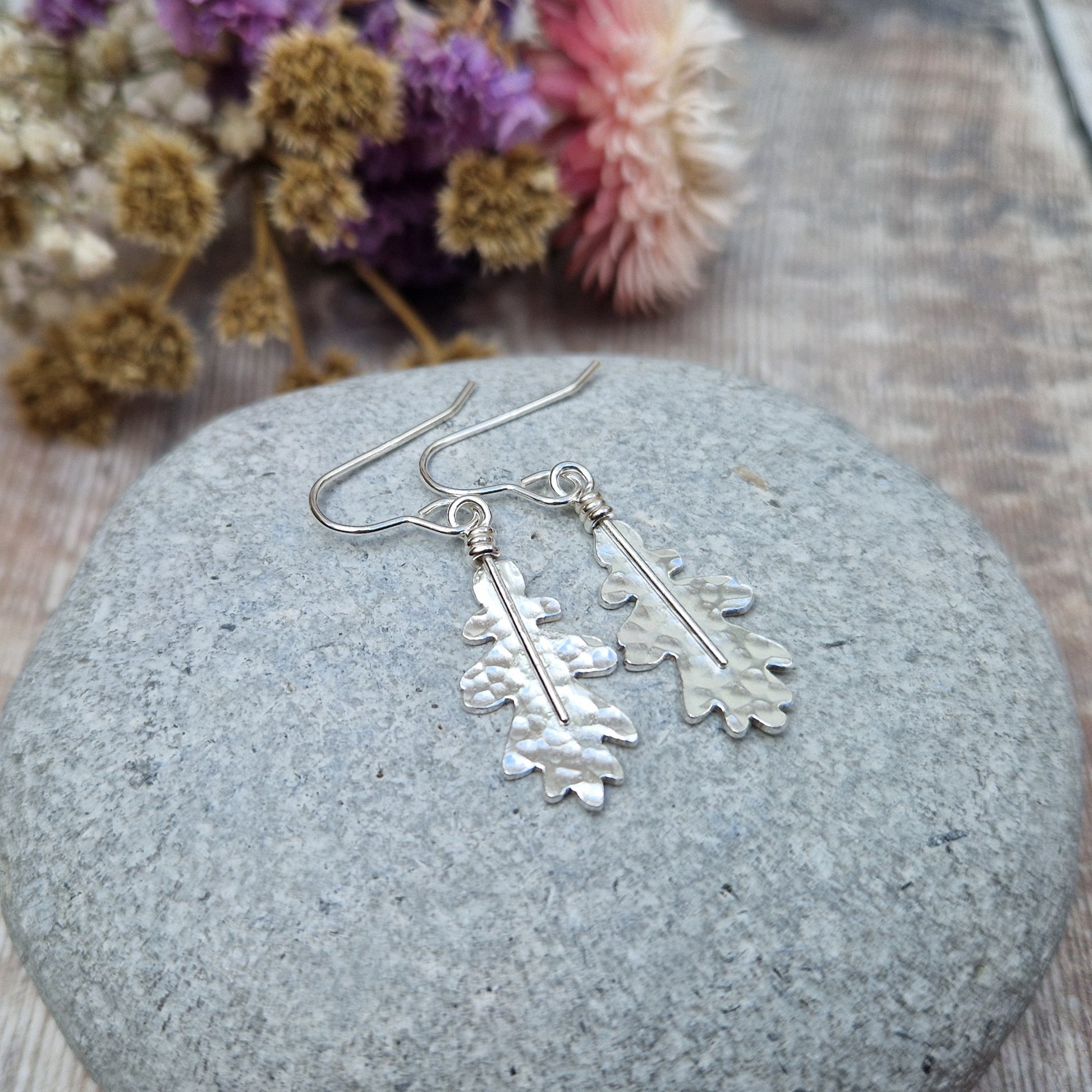 A close-up of sterling silver oak leaf earrings resting on a smooth grey stone. The hammered texture and organic shape of the leaves are highlighted, with soft-focus dried flowers in the background.