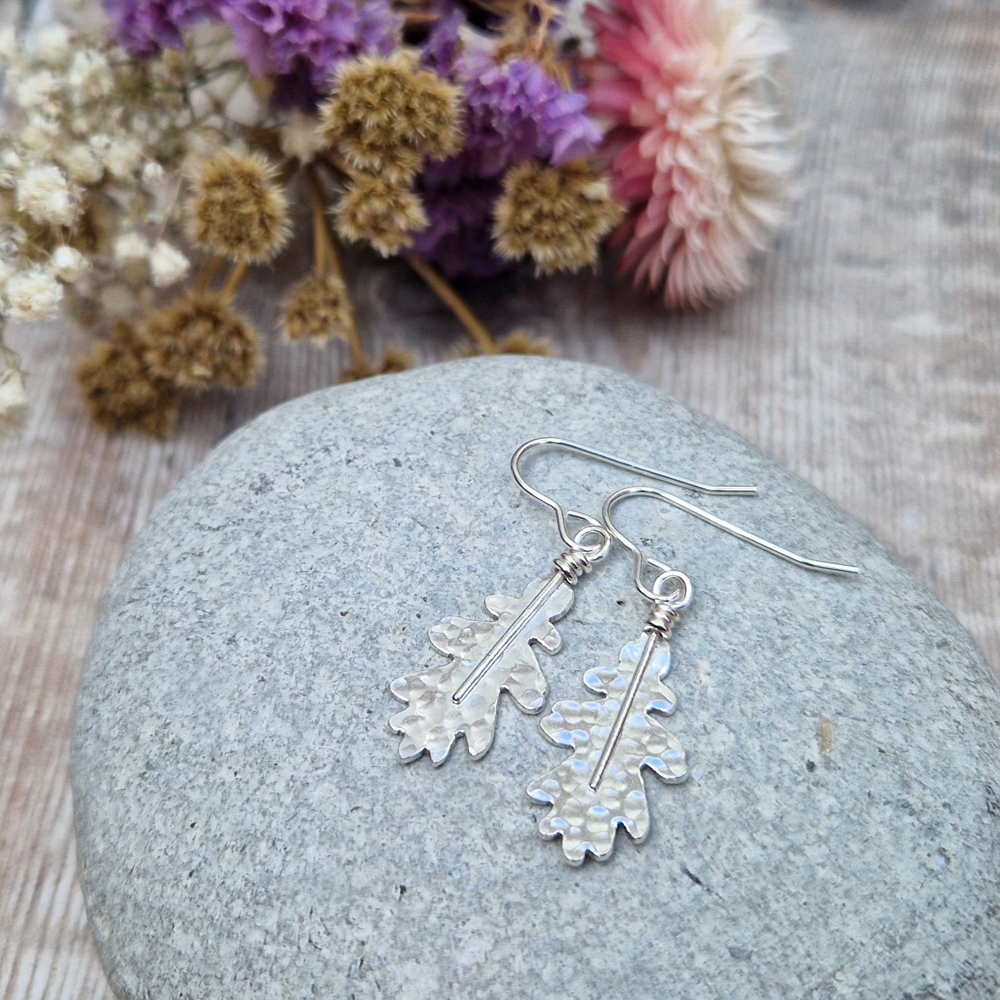 A close-up of sterling silver oak leaf earrings resting on a smooth grey stone. The hammered texture and organic shape of the leaves are highlighted, with soft-focus dried flowers in the background.