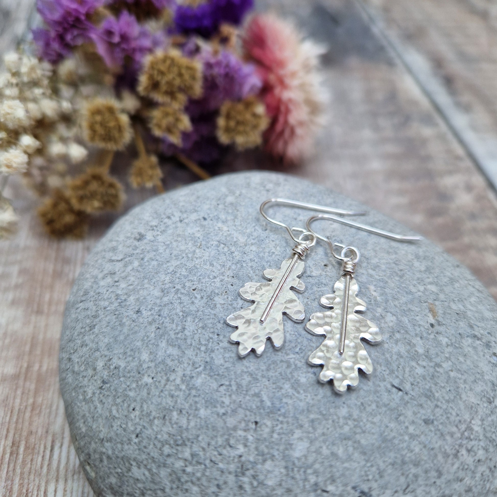 A close-up of sterling silver oak leaf earrings resting on a smooth grey stone. The hammered texture and organic shape of the leaves are highlighted, with soft-focus dried flowers in the background.