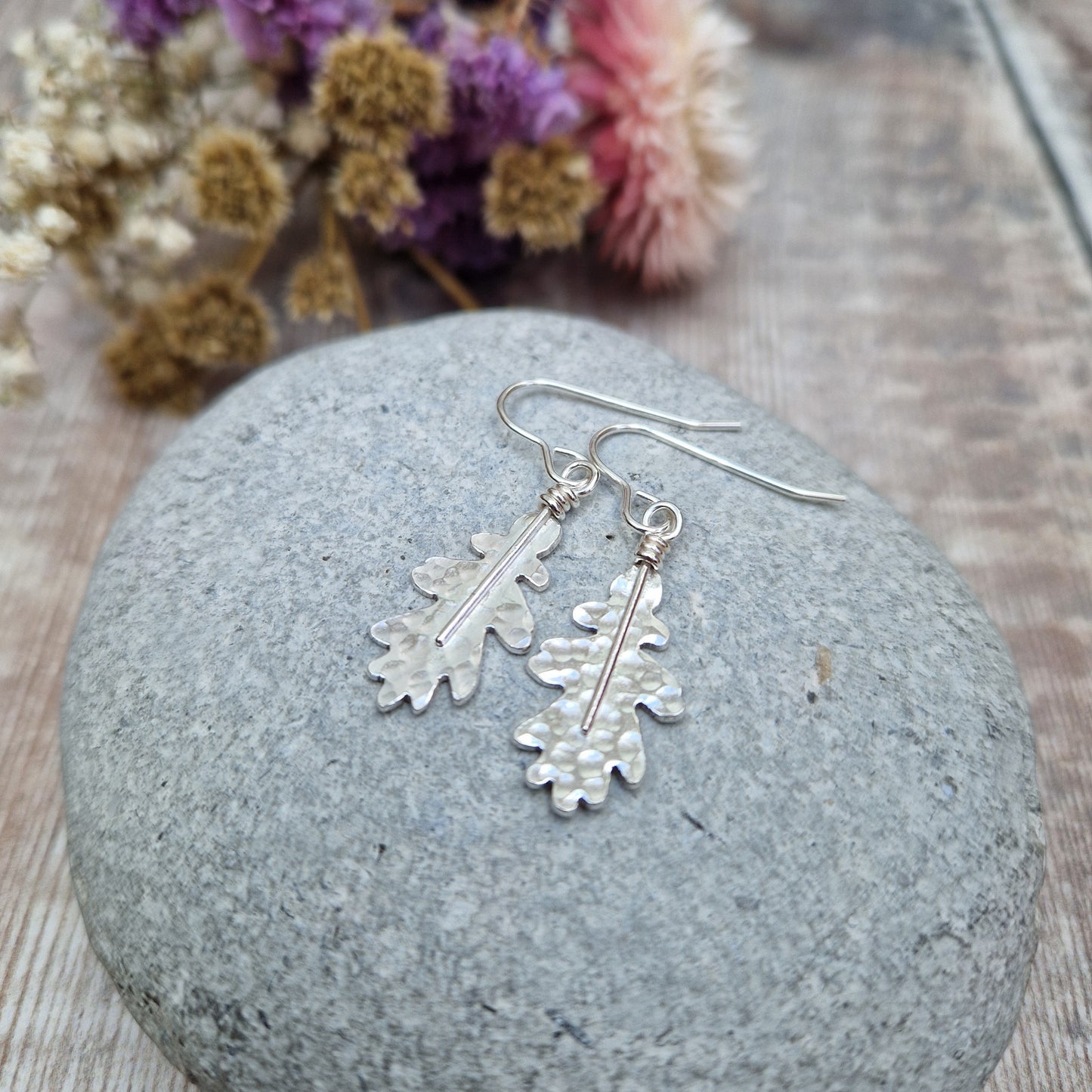 A close-up of sterling silver oak leaf earrings resting on a smooth grey stone. The hammered texture and organic shape of the leaves are highlighted, with soft-focus dried flowers in the background.