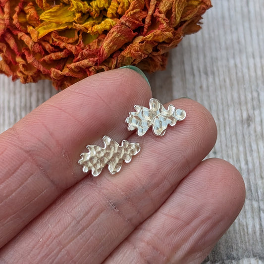 A pair of sterling silver oak leaf stud earrings with a hammered finish displayed on the fingertips of a hand, with a dried flower in the background.