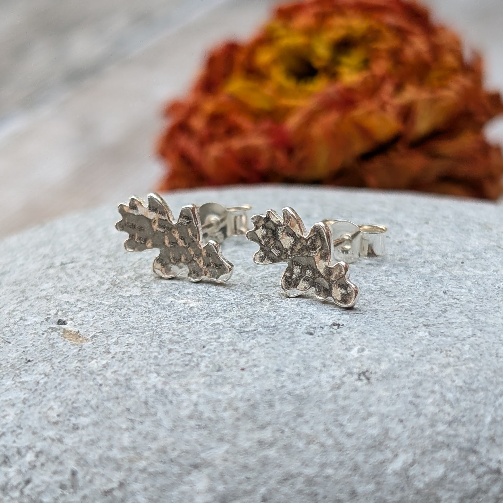 Close-up of a pair of sterling silver oak leaf stud earrings with a hammered finish, resting on a smooth grey stone with a blurred autumnal flower in the background.