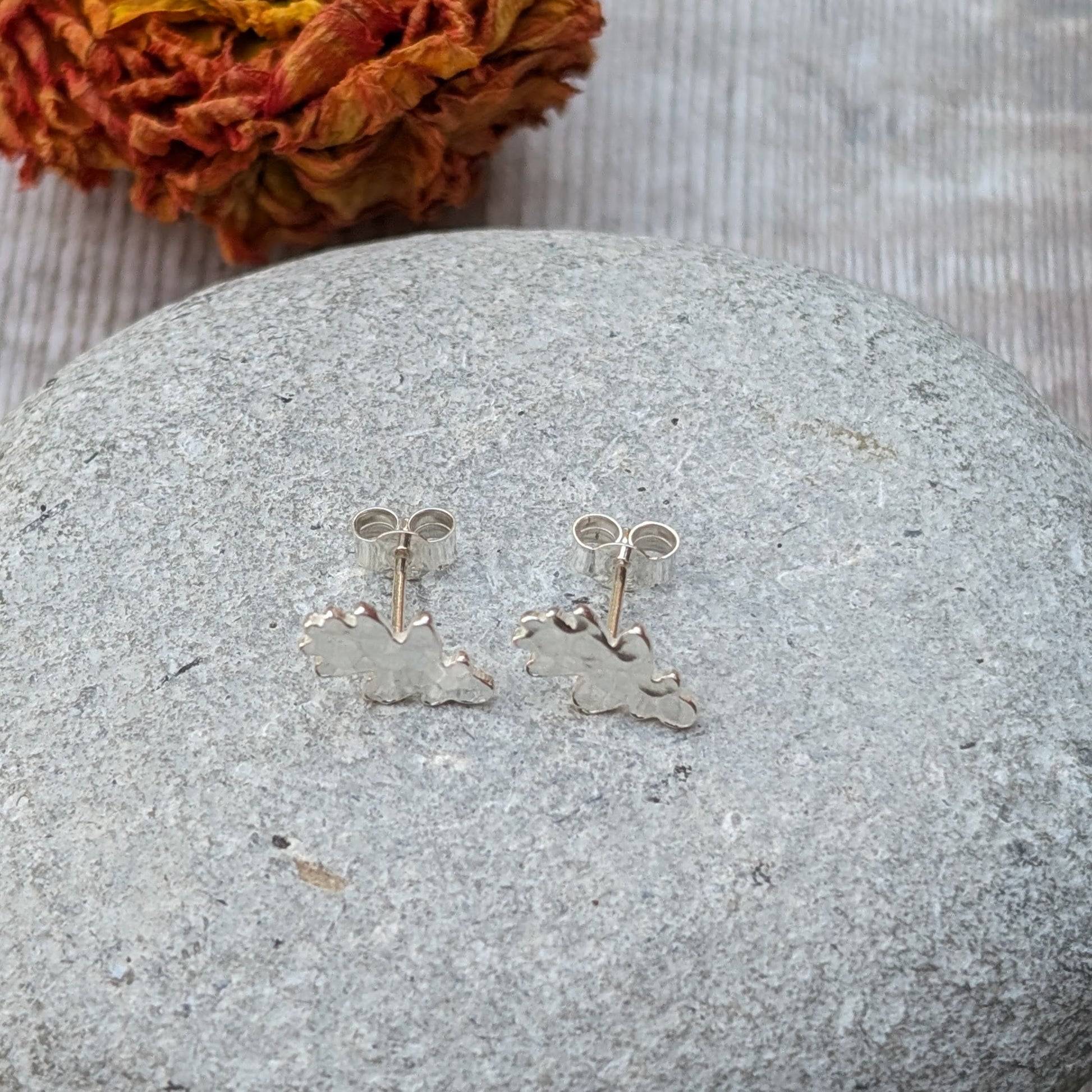 Close-up of a pair of sterling silver oak leaf stud earrings with a hammered finish, resting on a smooth grey stone with a blurred autumnal flower in the background.