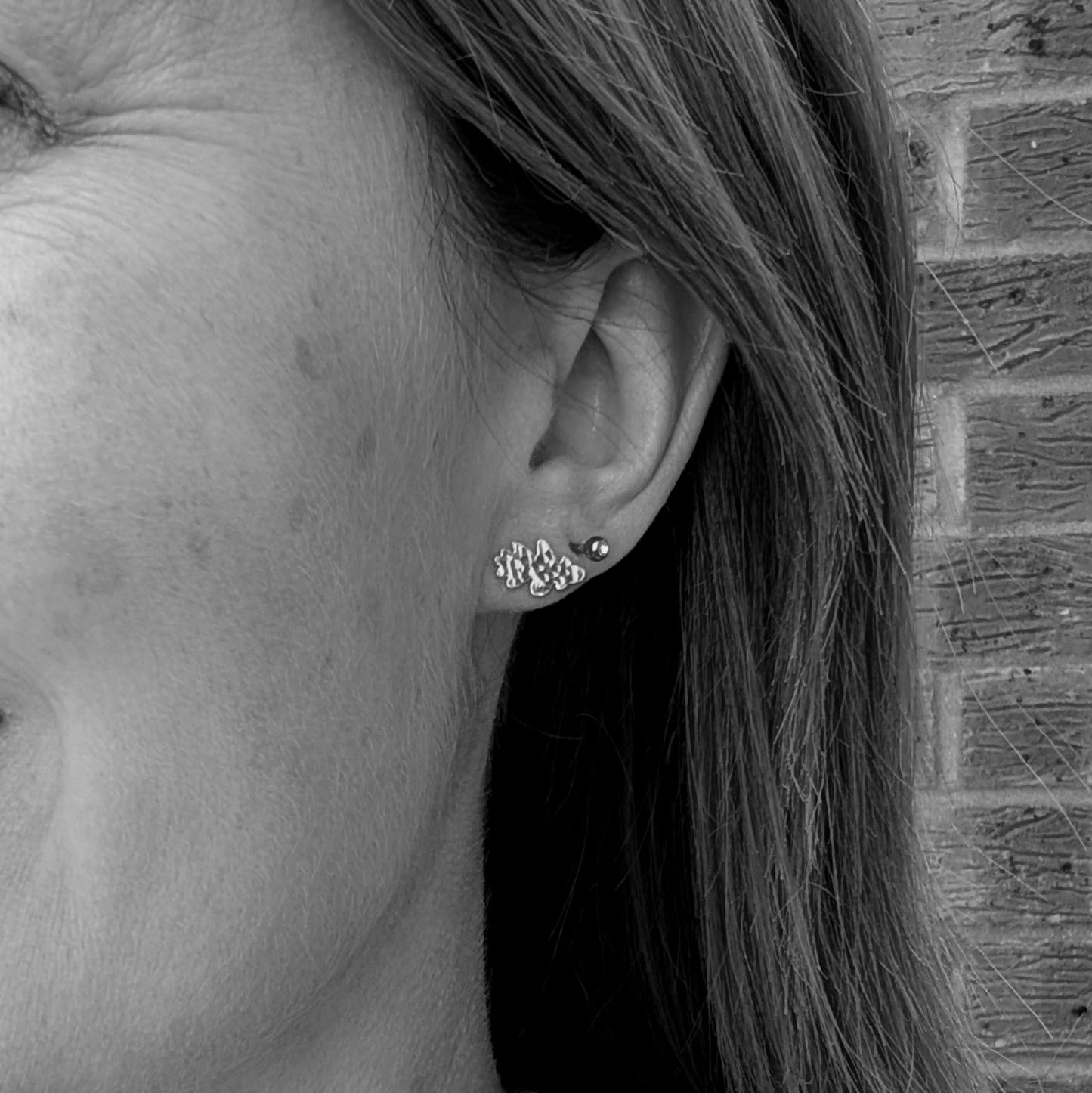 Black and white close-up of a person's ear wearing a sterling silver oak leaf stud earring with a hammered texture. Another smaller stud is visible in the second piercing.