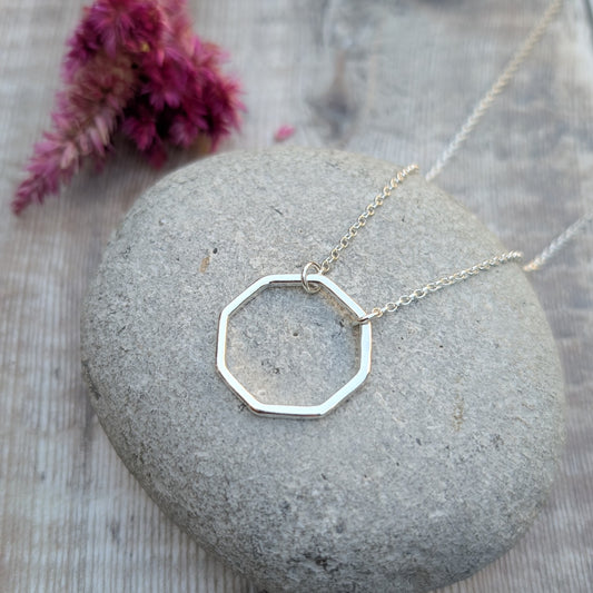 A sterling silver octagon pendant necklace resting on a smooth stone, with a blurred pink flower in the background.