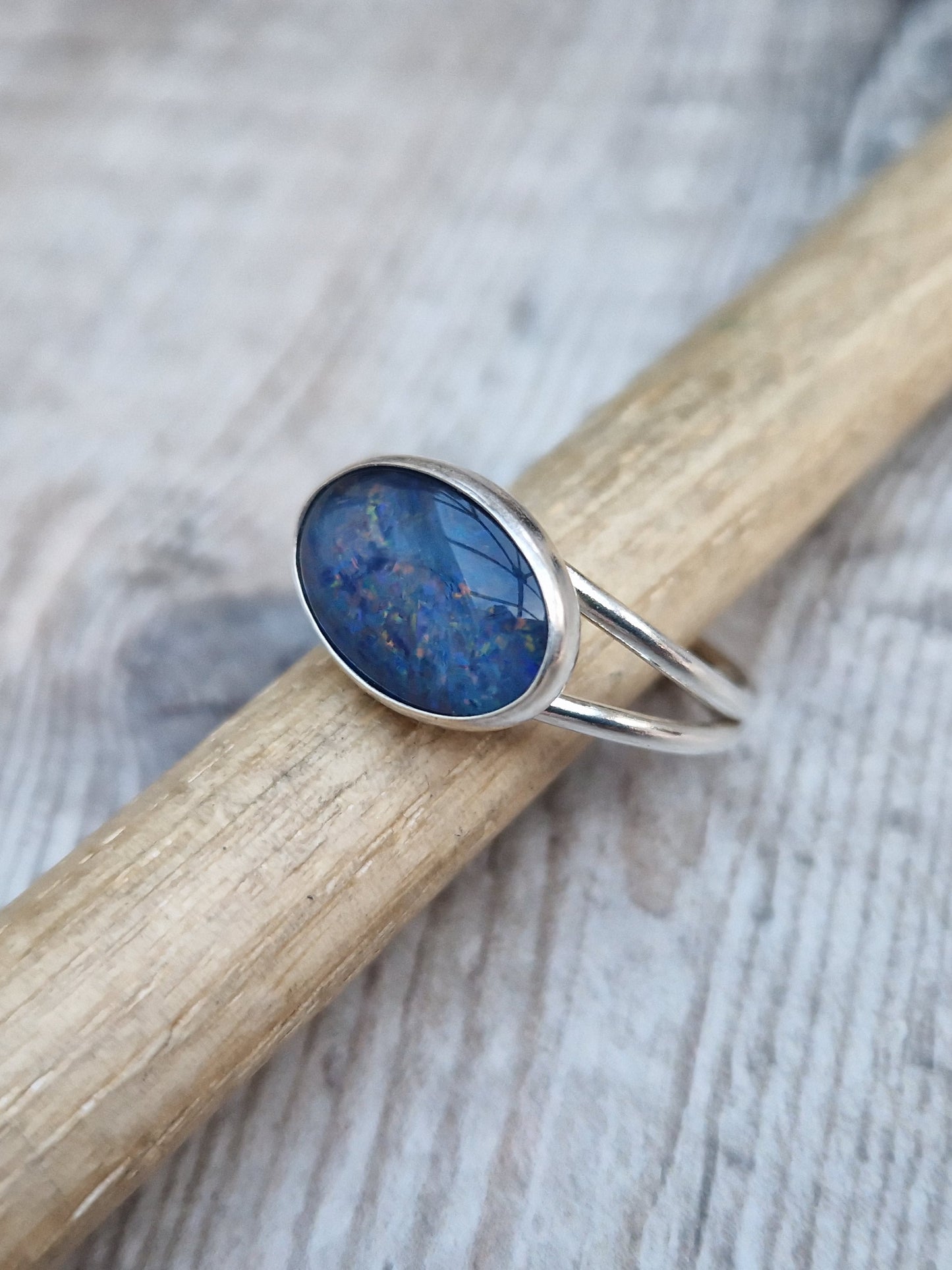 Blue Opal gemstone ring UK Size T ½ oval shape. Midnight blue stone with small flecks of green and orange, set in simple silver bezel surround and mounted lengthways on two silver bands that split at front and come together at back.