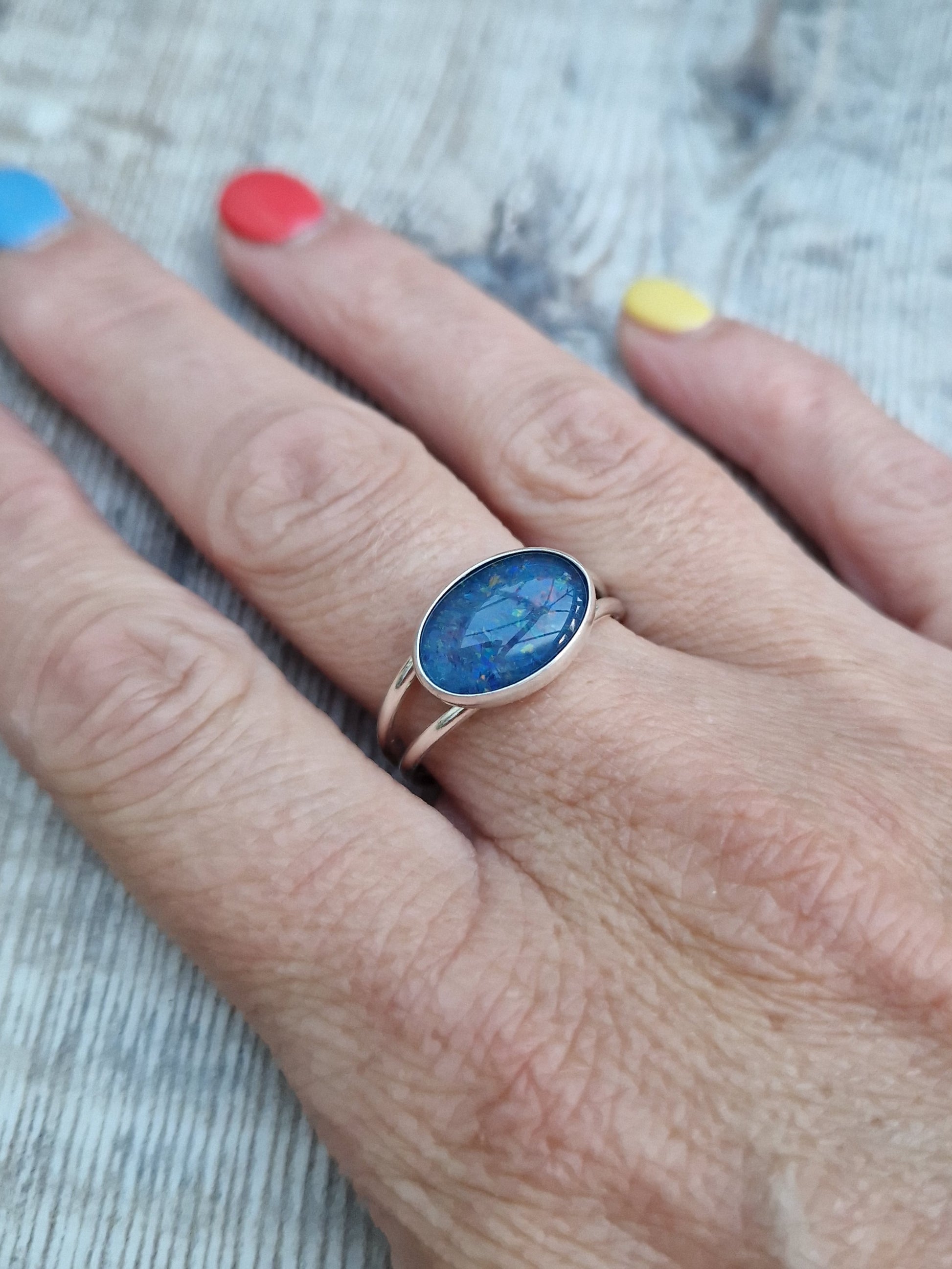 Blue Opal gemstone ring UK Size T ½ oval shape. Midnight blue stone with small flecks of green and orange, set in simple silver bezel surround and mounted lengthways on two silver bands that split at front and come together at back.