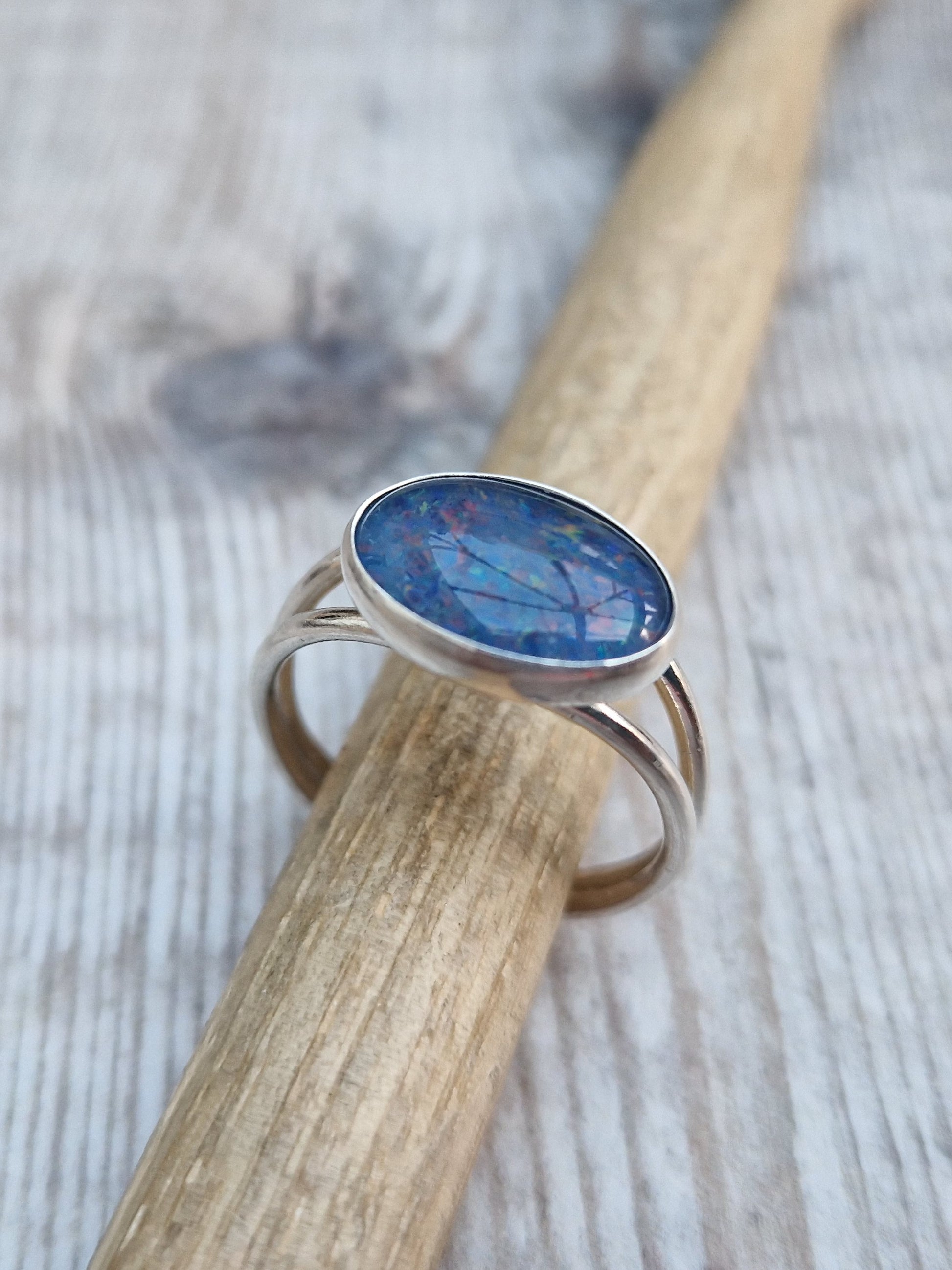 Blue Opal gemstone ring UK Size T ½ oval shape. Midnight blue stone with small flecks of green and orange, set in simple silver bezel surround and mounted lengthways on two silver bands that split at front and come together at back.