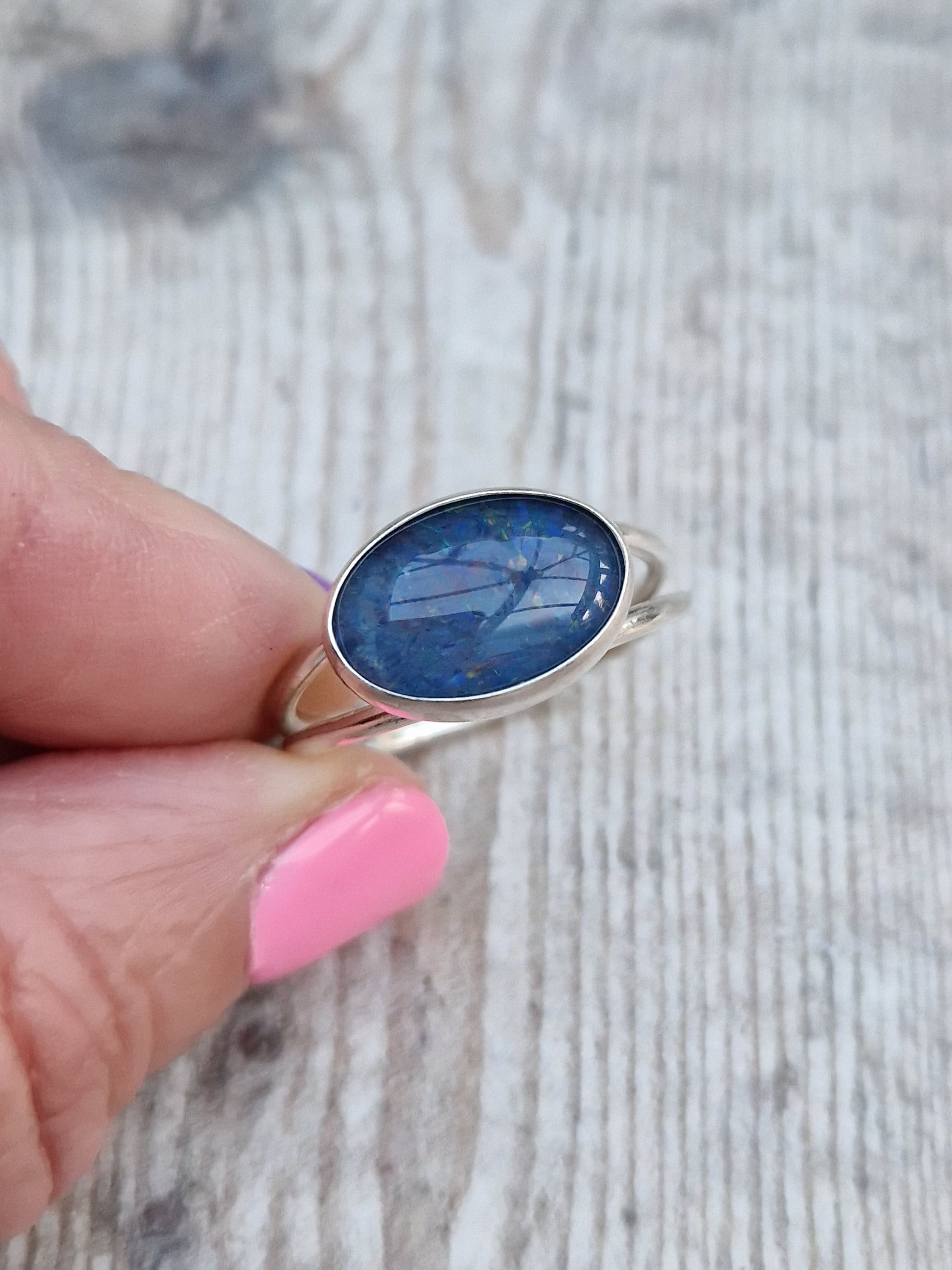 Blue Opal gemstone ring UK Size T ½ oval shape. Midnight blue stone with small flecks of green and orange, set in simple silver bezel surround and mounted lengthways on two silver bands that split at front and come together at back.