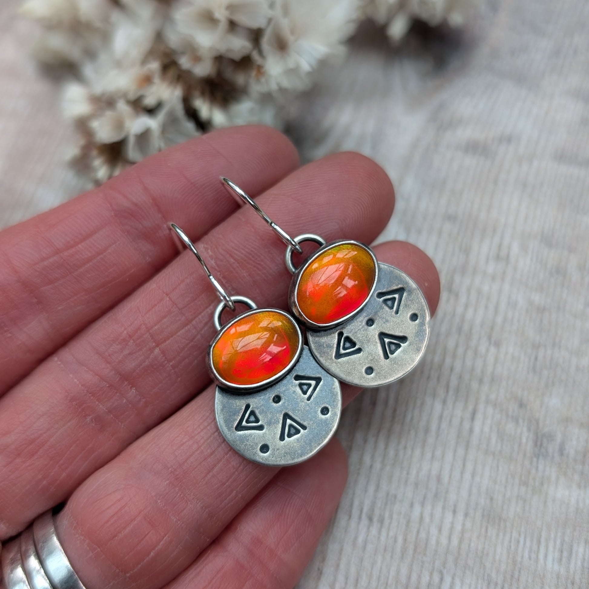 Each earring features one oval-shaped orange opal gemstone with vibrant hues set into an oxidised silver bezel mounted onto a round silver disc with geometric patterns, including small triangles and dots suspended from silver earring wire.