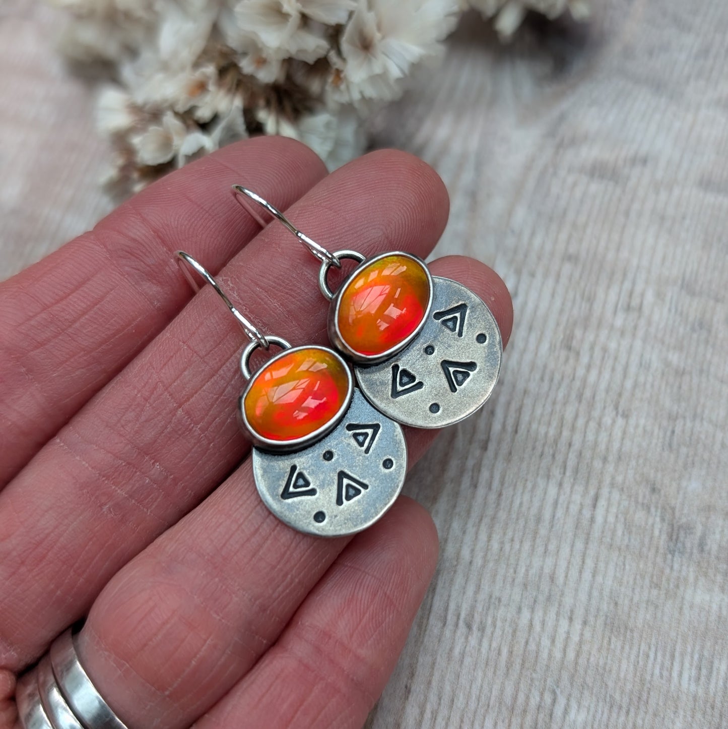 Each earring features one oval-shaped orange opal gemstone with vibrant hues set into an oxidised silver bezel mounted onto a round silver disc with geometric patterns, including small triangles and dots suspended from silver earring wire.