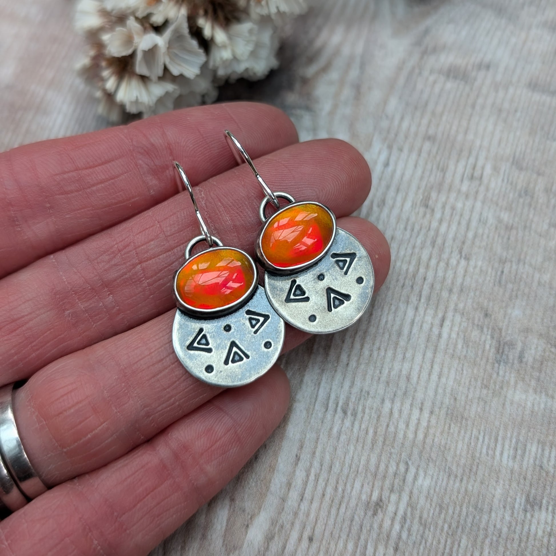 Each earring features one oval-shaped orange opal gemstone with vibrant hues set into an oxidised silver bezel mounted onto a round silver disc with geometric patterns, including small triangles and dots suspended from silver earring wire.