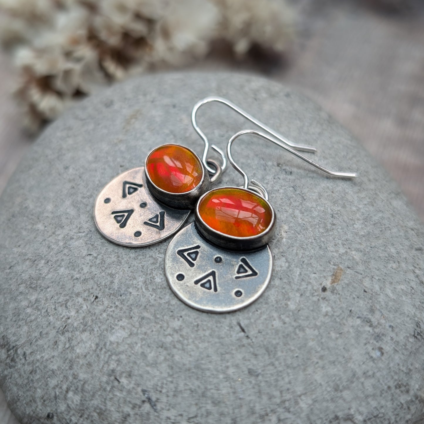 Each earring features one oval-shaped orange opal gemstone with vibrant hues set into an oxidised silver bezel mounted onto a round silver disc with geometric patterns, including small triangles and dots suspended from silver earring wire.