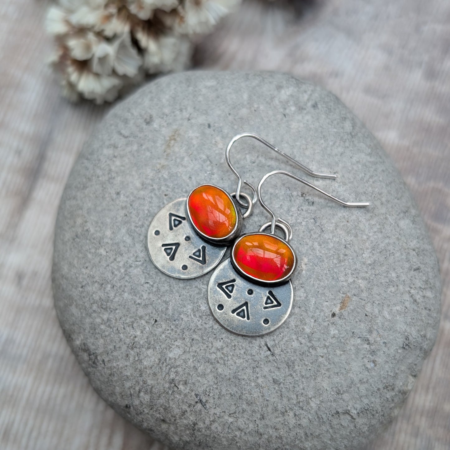 Each earring features one oval-shaped orange opal gemstone with vibrant hues set into an oxidised silver bezel mounted onto a round silver disc with geometric patterns, including small triangles and dots suspended from silver earring wire.