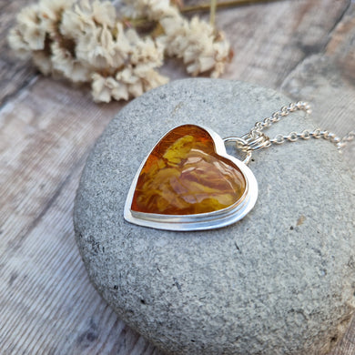Surfite Orange heart shaped pendant set in Sterling Silver bezel surround, attached to a silver chain via two small circle links. The orange Surfite has orange, gold, brown and yellow flecks running through it with a smooth, gloss finish. The rear of the bezel has been left open to view the surfite with pink hued flecks.