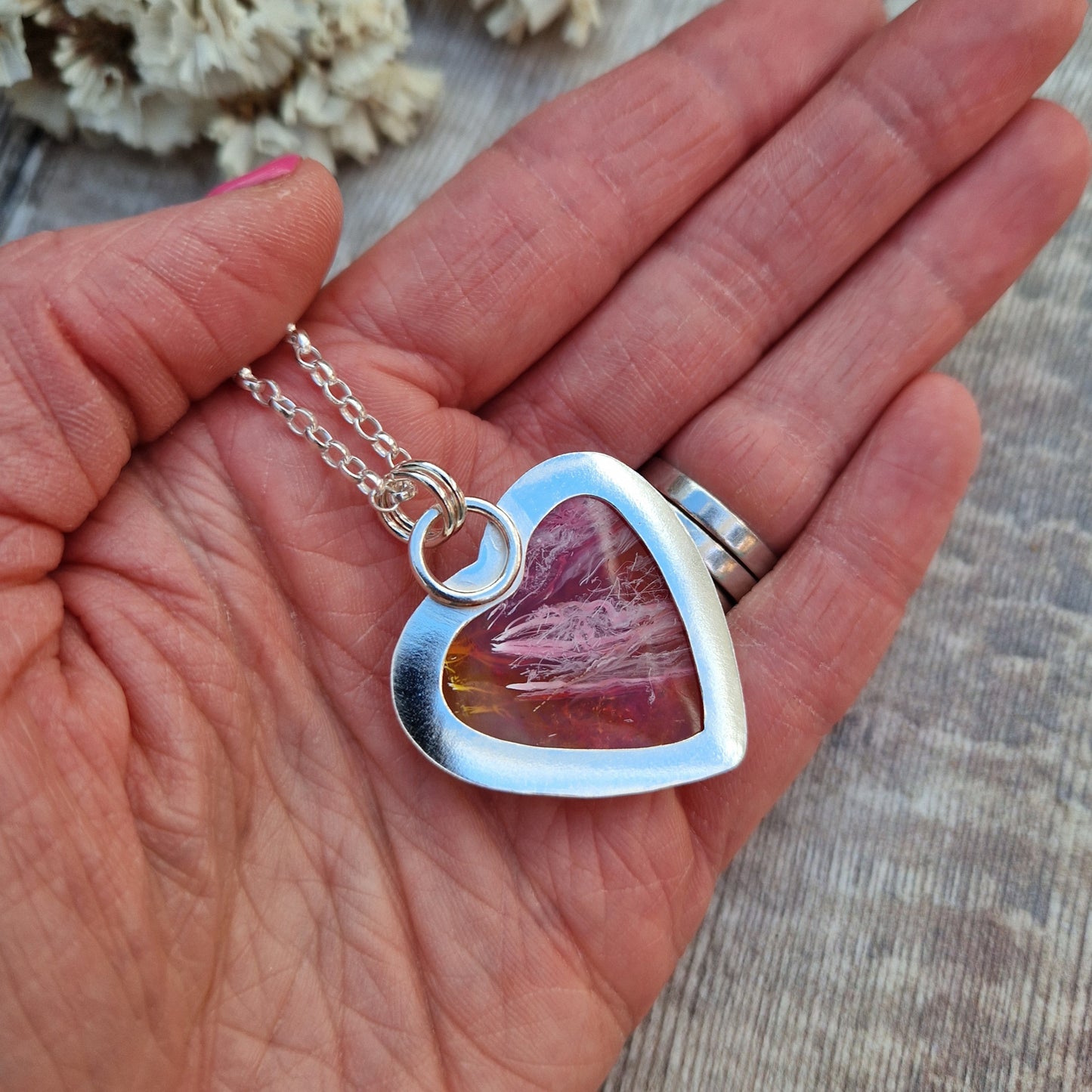 Surfite Orange heart shaped pendant set in Sterling Silver bezel surround, attached to a silver chain via two small circle links. The orange Surfite has orange, gold, brown and yellow flecks running through it with a smooth, gloss finish. The rear of the bezel has been left open to view the surfite with pink hued flecks.