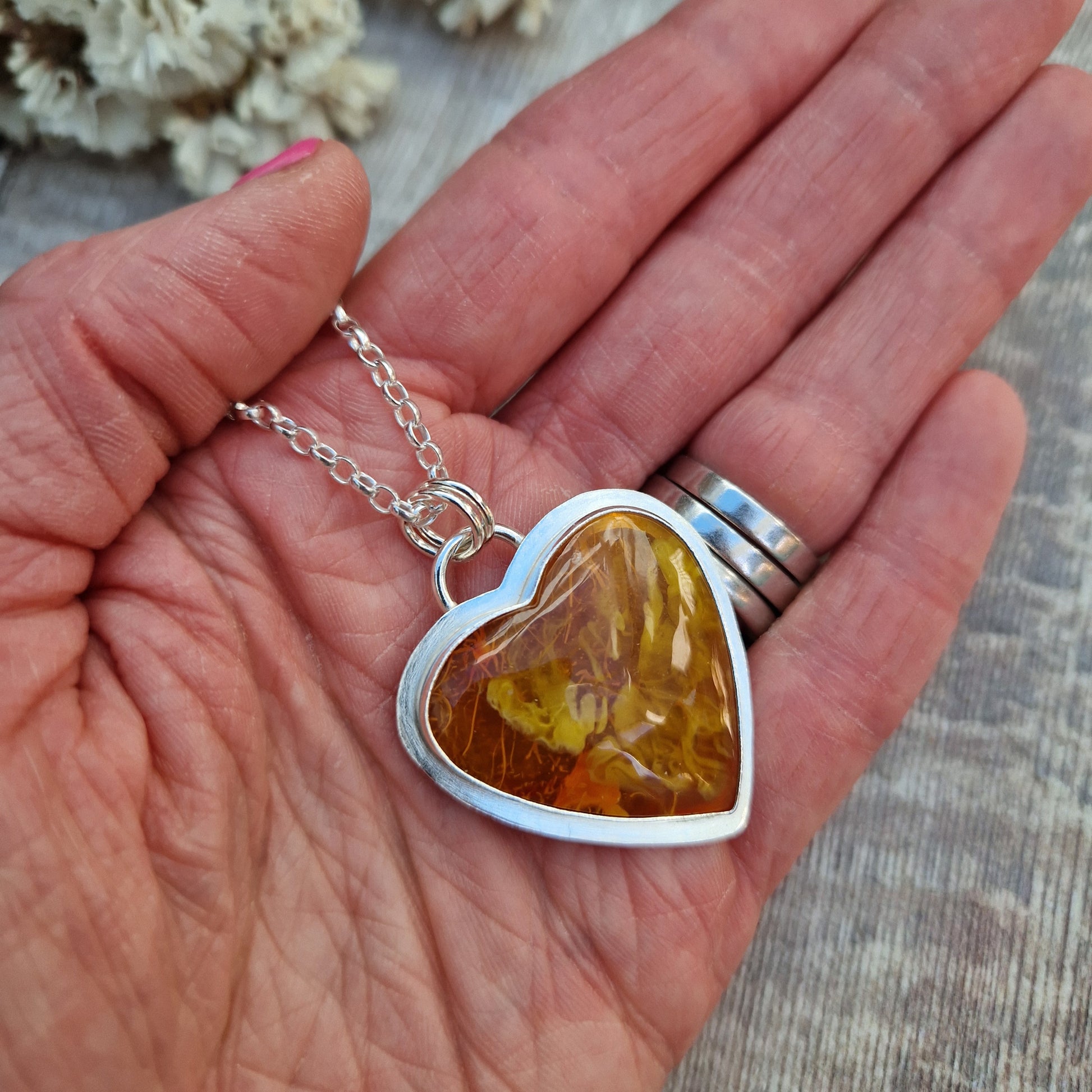 Surfite Orange heart shaped pendant set in Sterling Silver bezel surround, attached to a silver chain via two small circle links. The orange Surfite has orange, gold, brown and yellow flecks running through it with a smooth, gloss finish. The rear of the bezel has been left open to view the surfite with pink hued flecks.