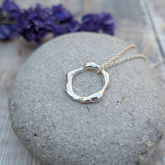 A close-up of a handmade recycled sterling silver organic circle necklace resting on a smooth stone, with a blurred background of purple flowers.