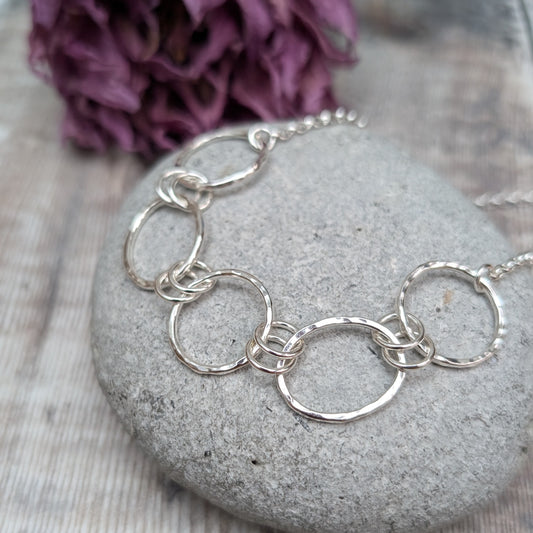 Five open silver circles with hammered finish linked with each other via two small silver circles in a repeating pattern. Each circle at each end of the pattern is attached to a silver chain. 