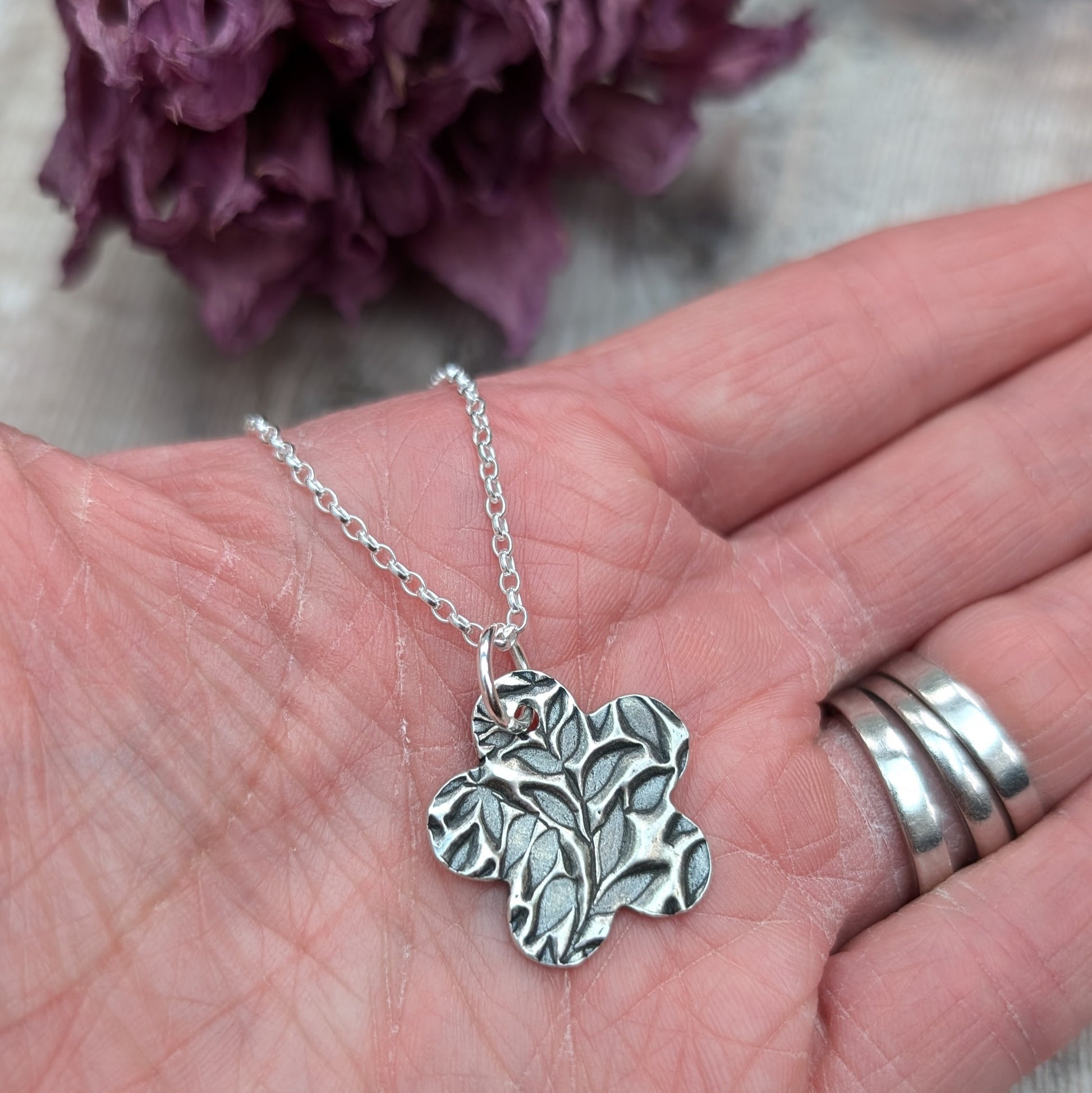 An oxidized silver pendant with a floral leaf pattern design held in a person's hand for size reference, with a wooden background.