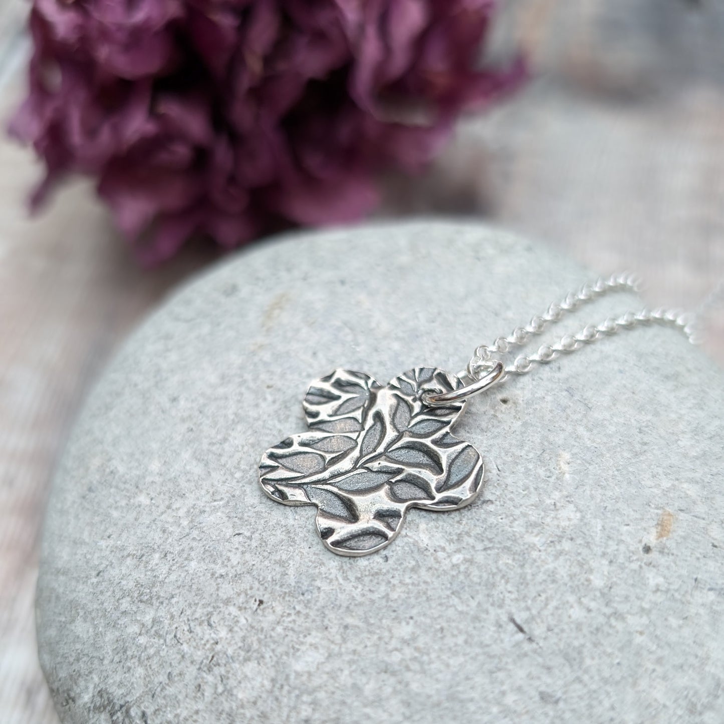 An oxidized silver pendant with a floral leaf pattern design on a smooth gray stone background. The pendant is on a silver chain.