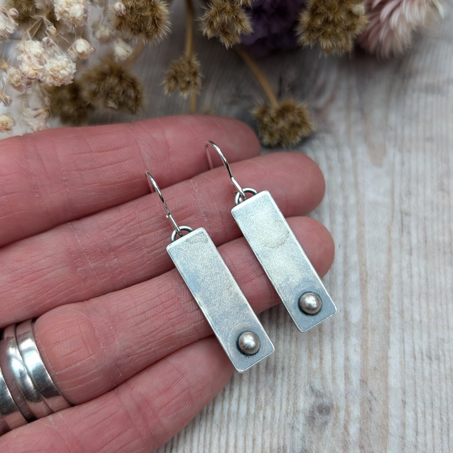 A pair of rectangular oxidized silver earrings with a small round silver pebble at the bottom of each rectangle, held in a person's hand for size reference, with a wooden background.